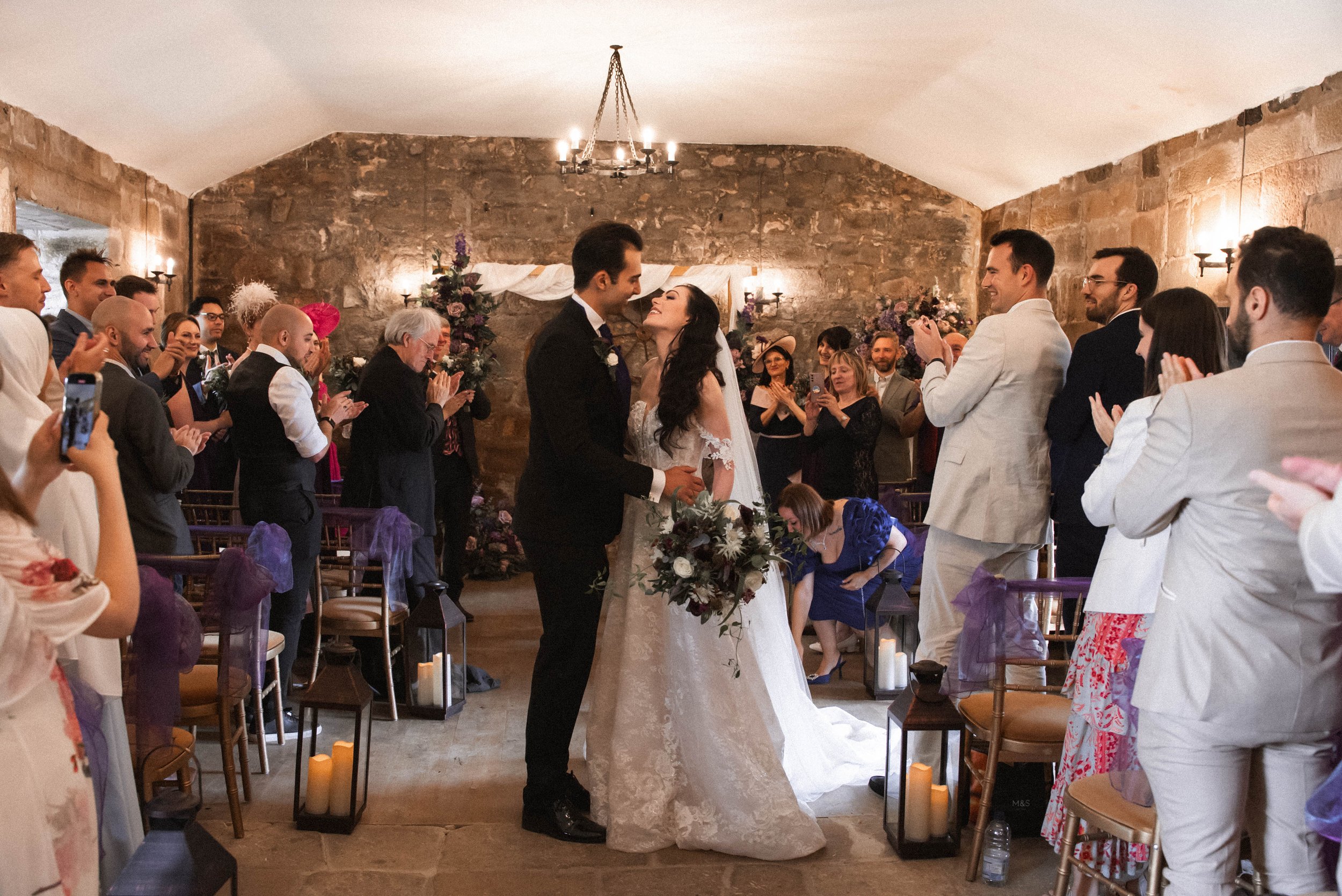 A wedding ceremony with a bride and groom holding hands at the altar, surrounded by friends and family in a rustic indoor setting with stone walls, decorated with flowers and candlelit candles.