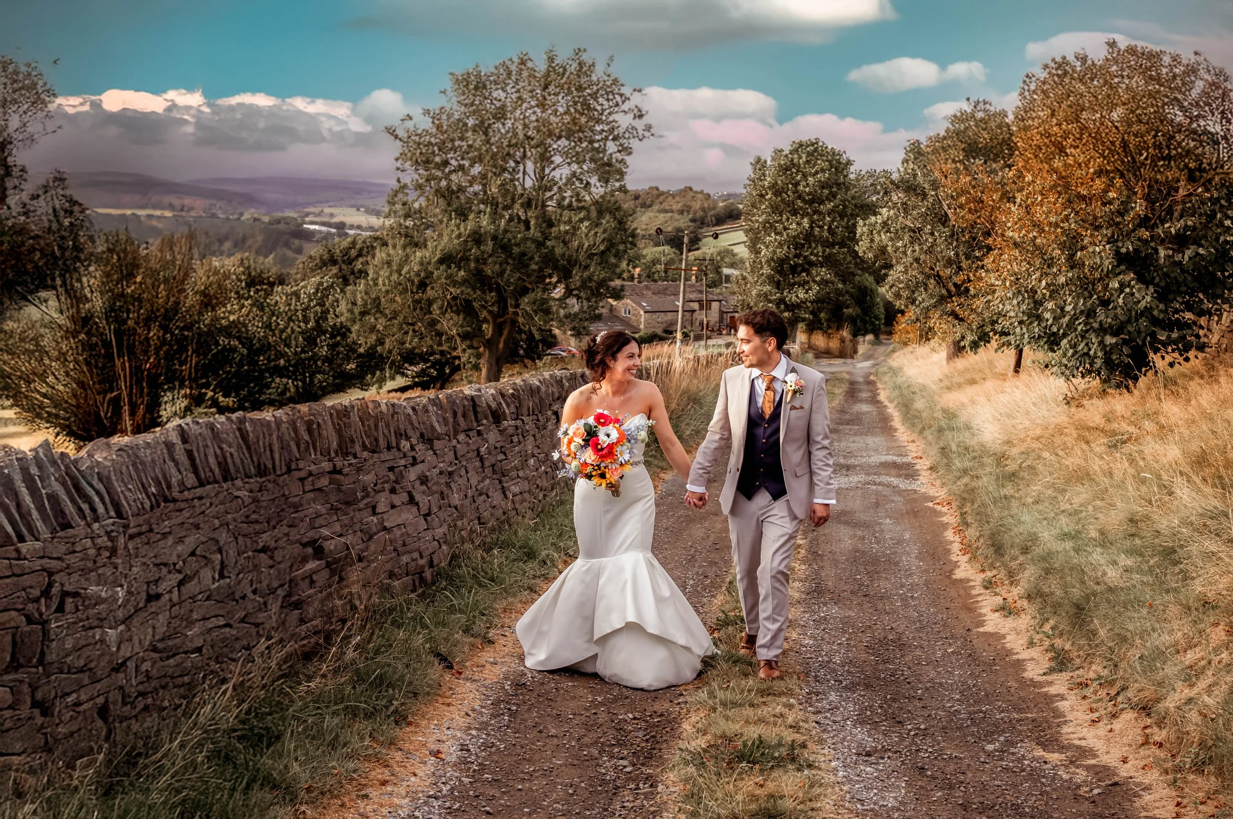 A newly married couple walking hand-in-hand on a rural dirt road, with trees and rolling hills in the background during sunset.