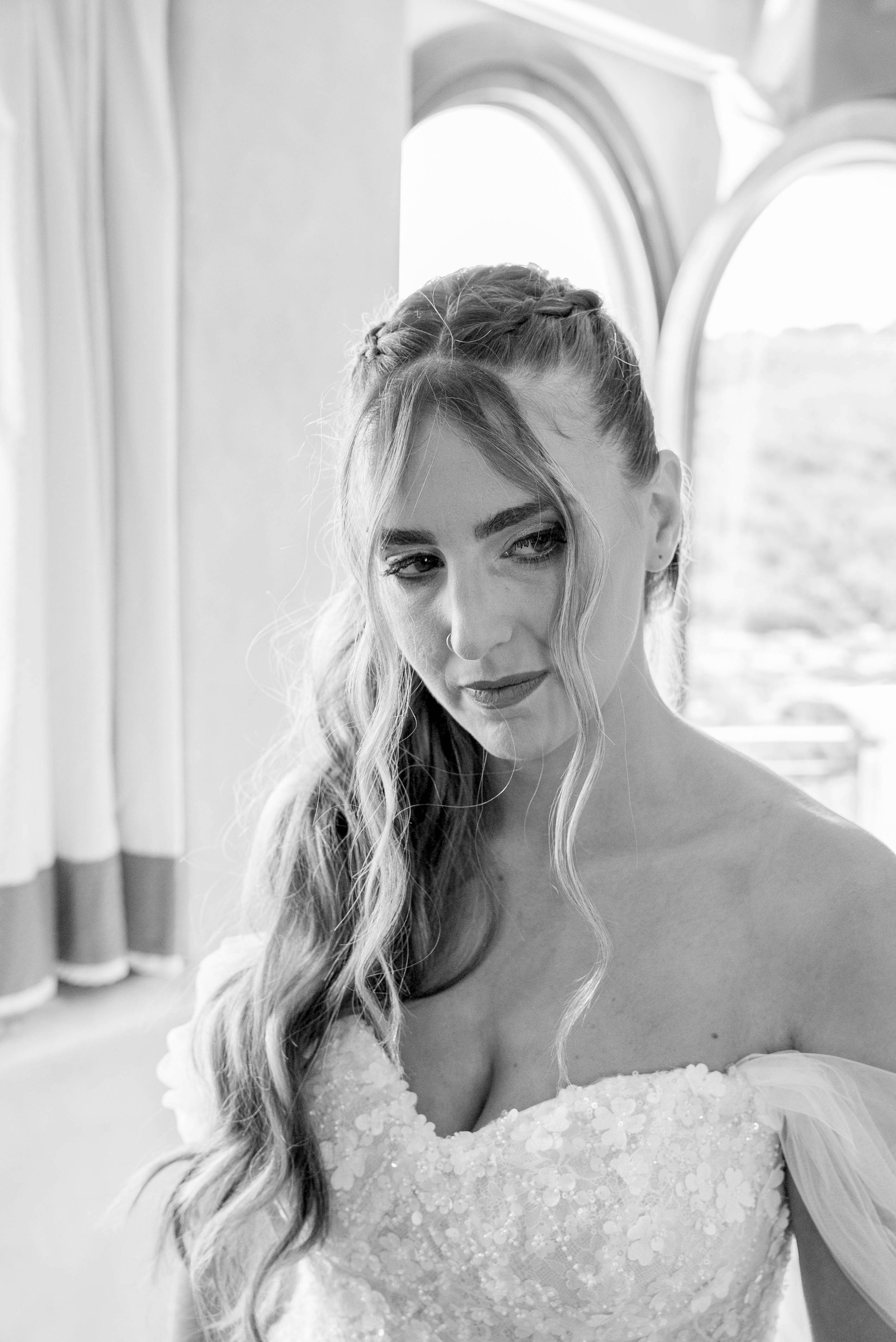 A black-and-white photo of a woman with long wavy hair in an updo, wearing a bridal gown with floral embellishments, looking down and to her left near a window in a well-lit room.