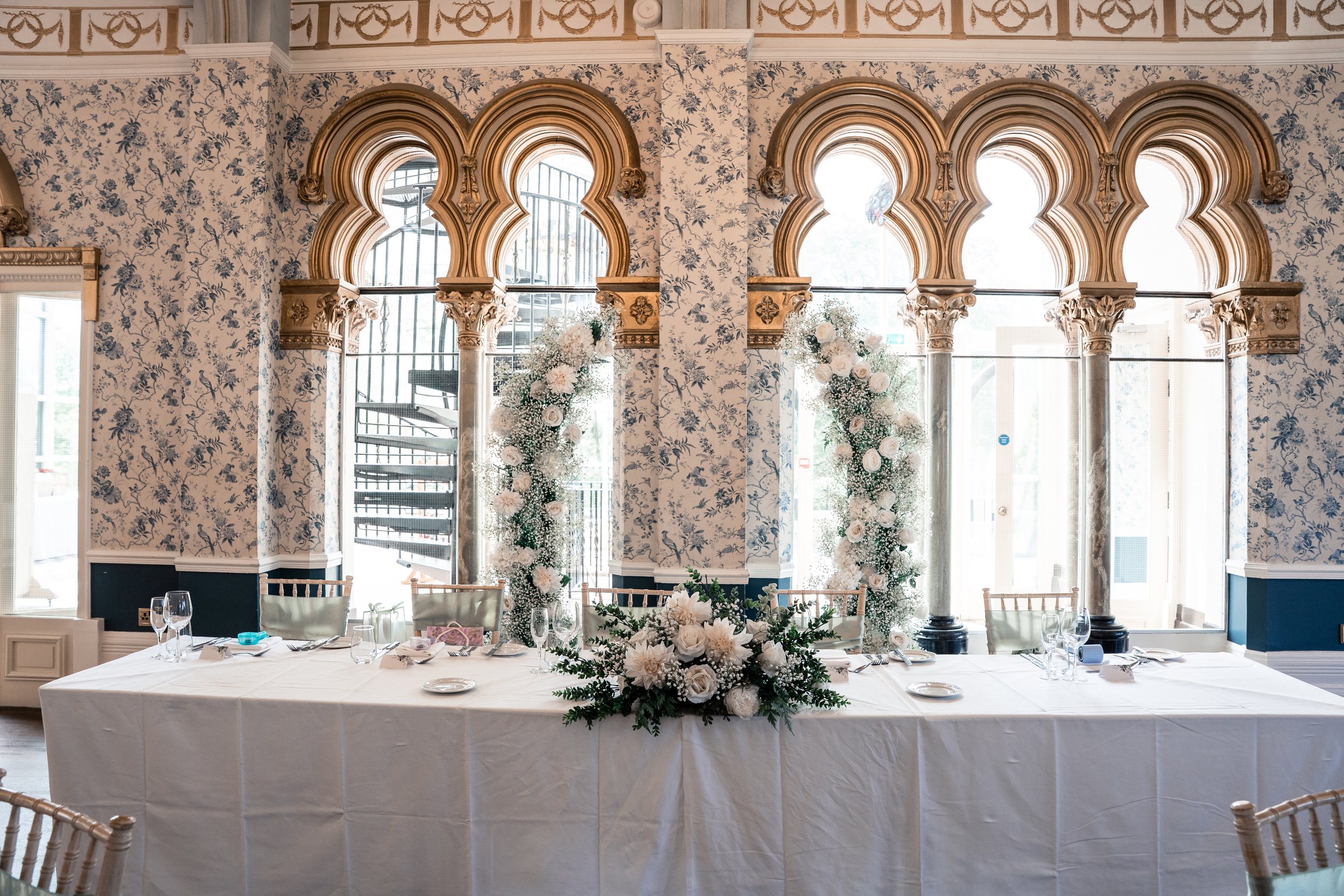 A decorated banquet table in a bright, ornate room with large arched windows and floral wallpaper, featuring floral arrangements and table settings for a formal event.