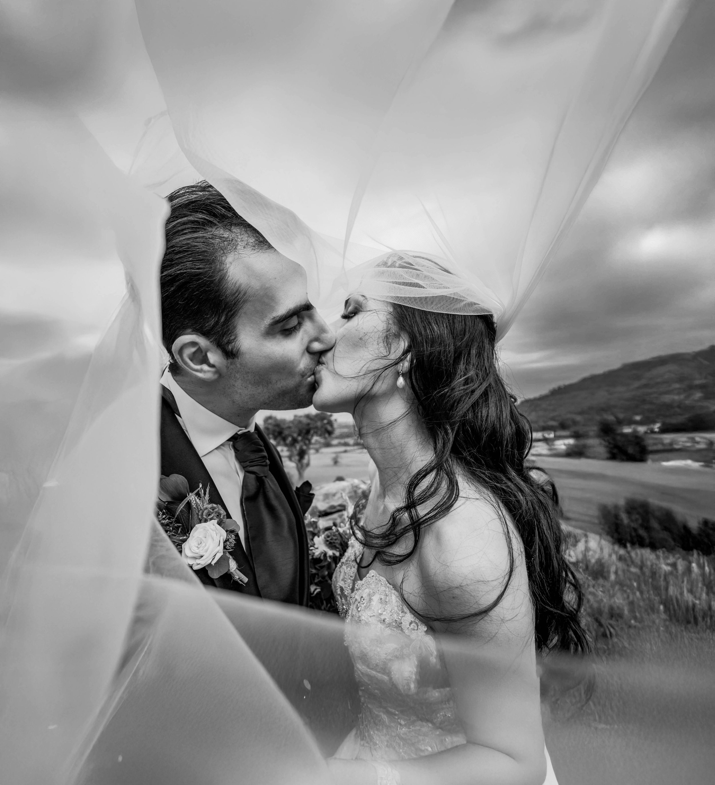 A black and white photograph of a bride and groom kissing outdoors, with a landscape of hills and fields in the background, under a cloudy sky.