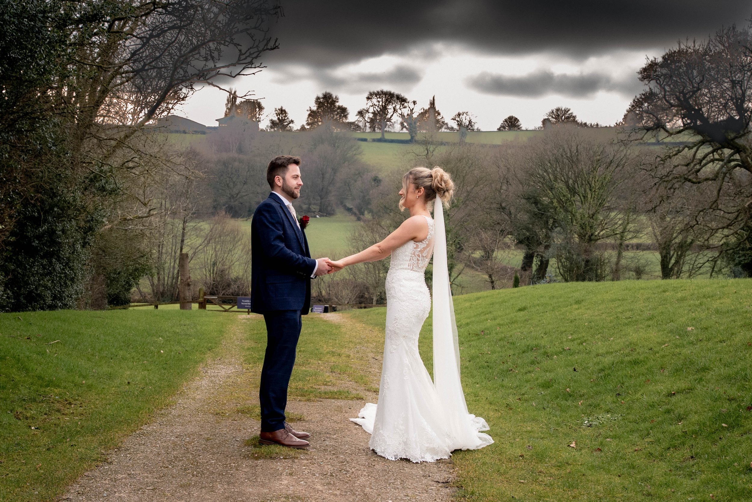 A couple dressed in wedding attire holding hands and facing each other outdoors on a cloudy day with trees and green hills in the background.