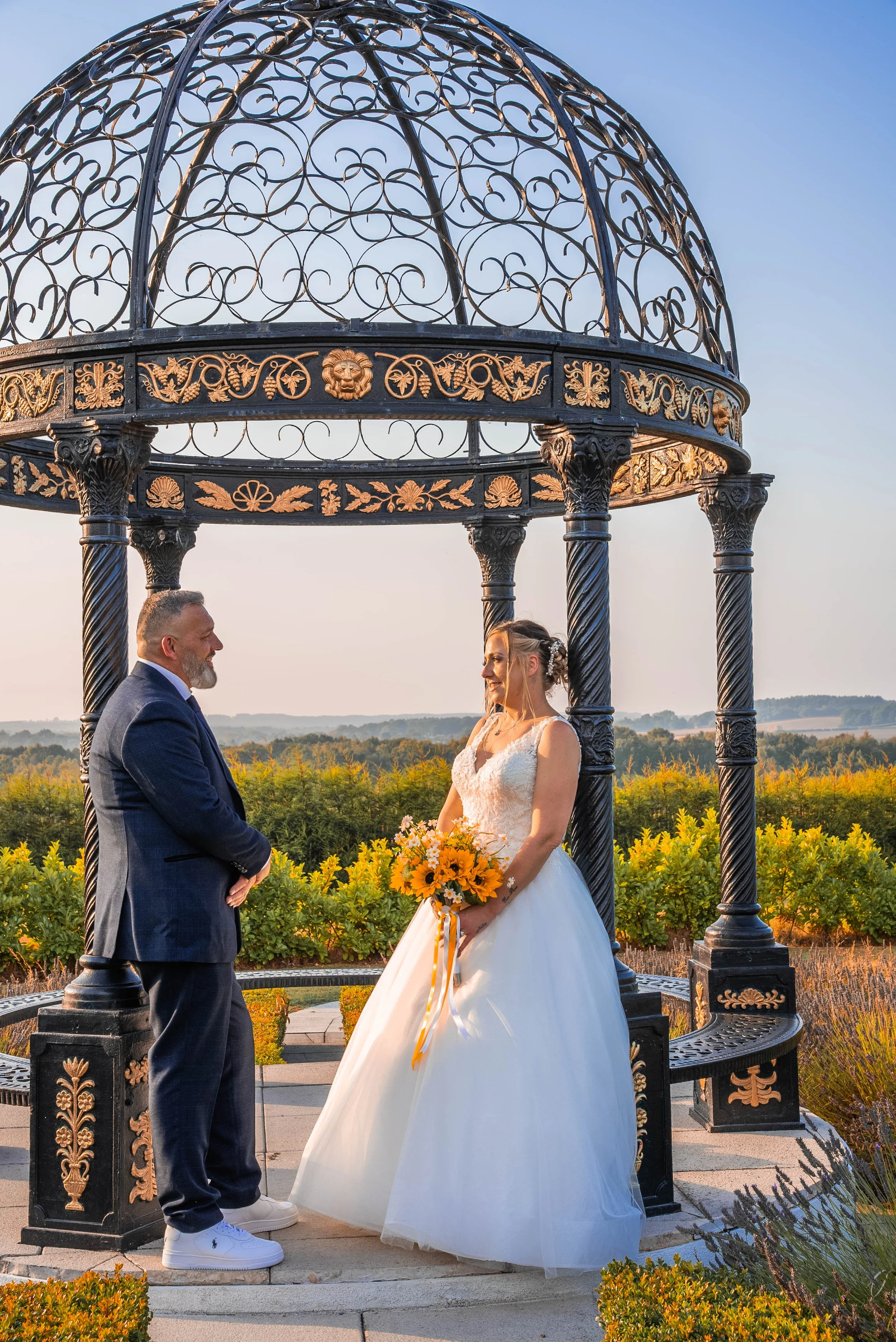 A bride in a white wedding dress holding a bouquet of sunflowers and another person in a dark suit standing in front of an ornate black and gold gazebo outdoors at sunset.