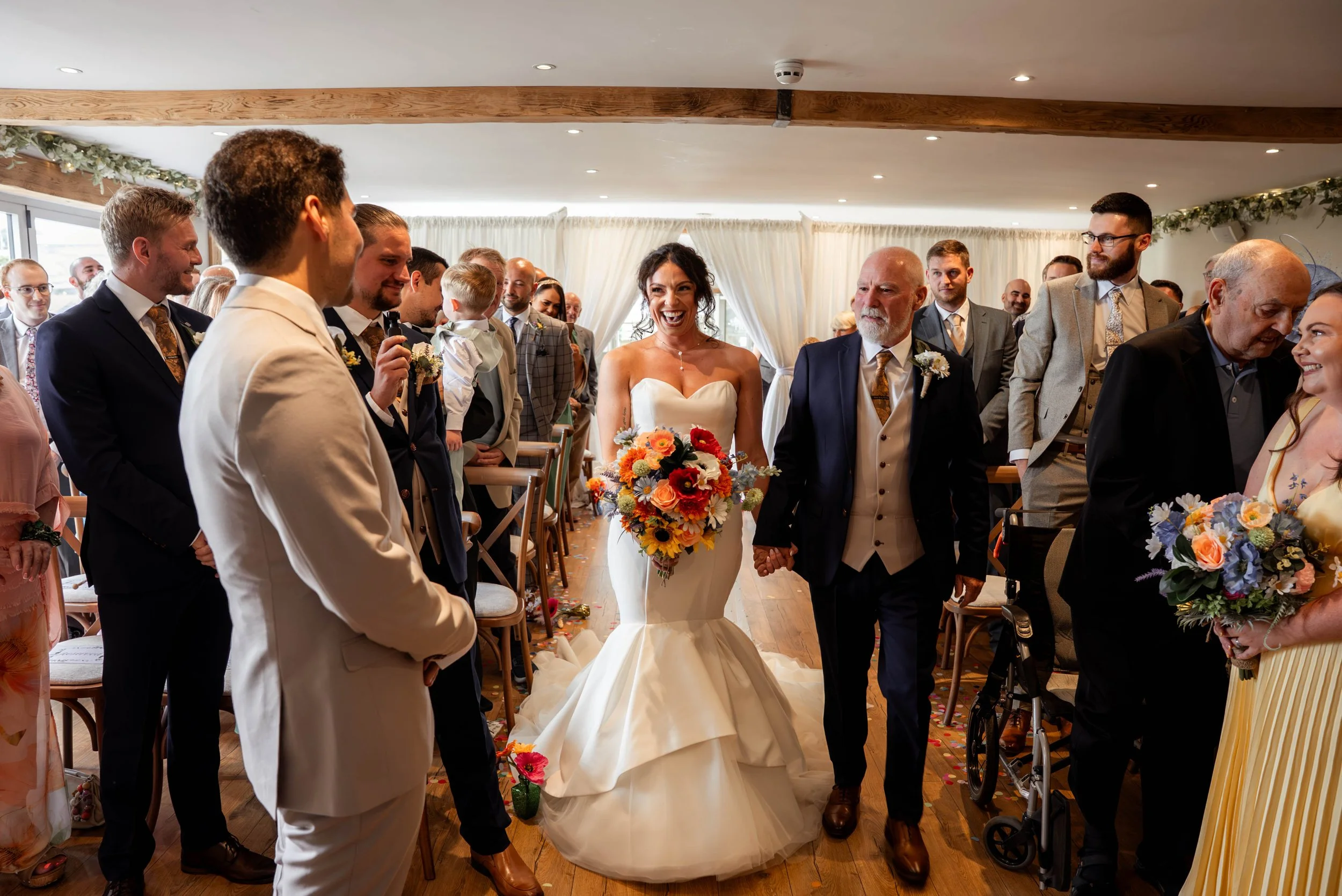A bride walking down the aisle with her father, holding a colorful bouquet, surrounded by wedding guests in an indoor ceremony.