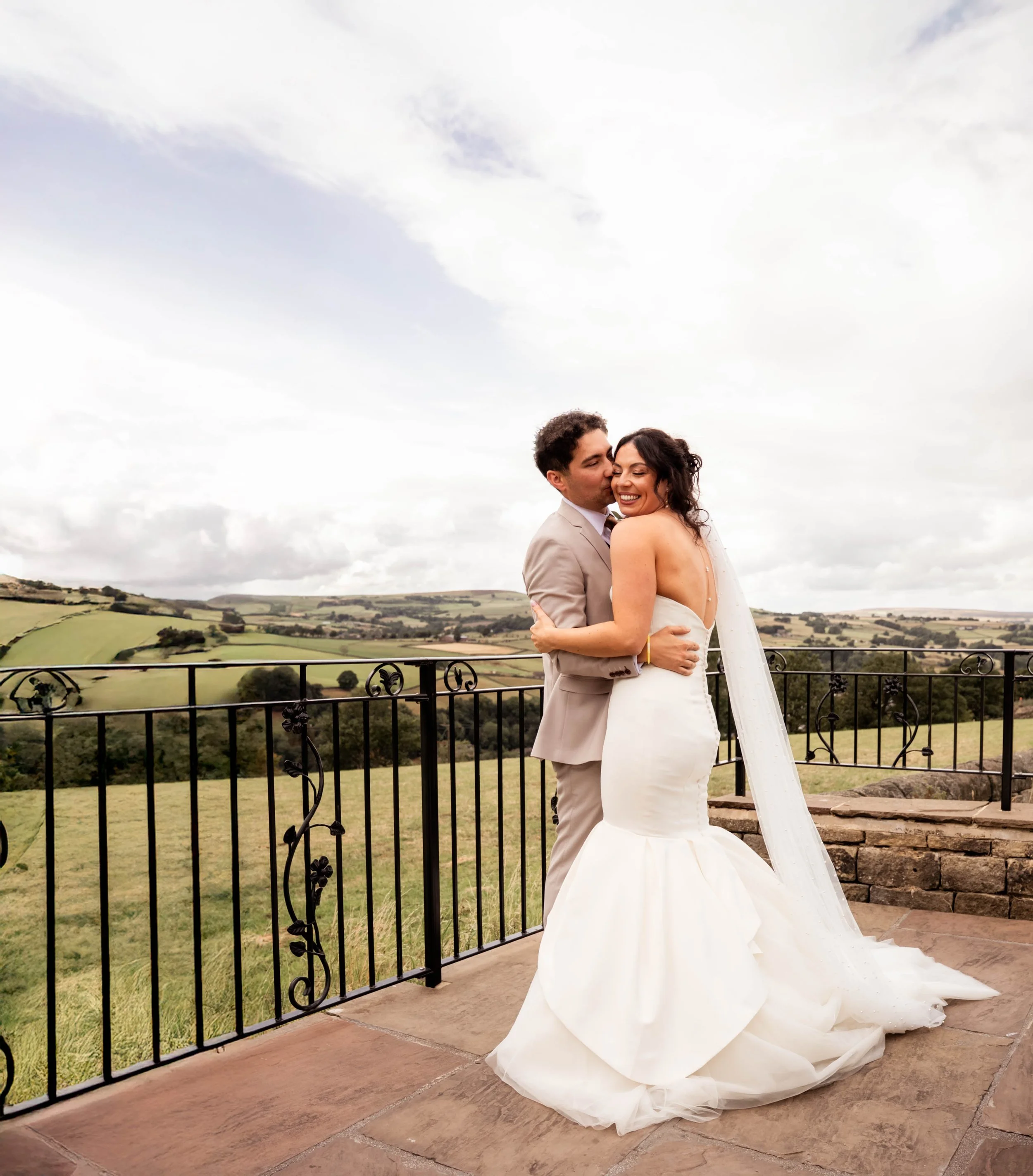 A newlywed couple embraces on a balcony overlooking a green countryside with hills under a cloudy sky.