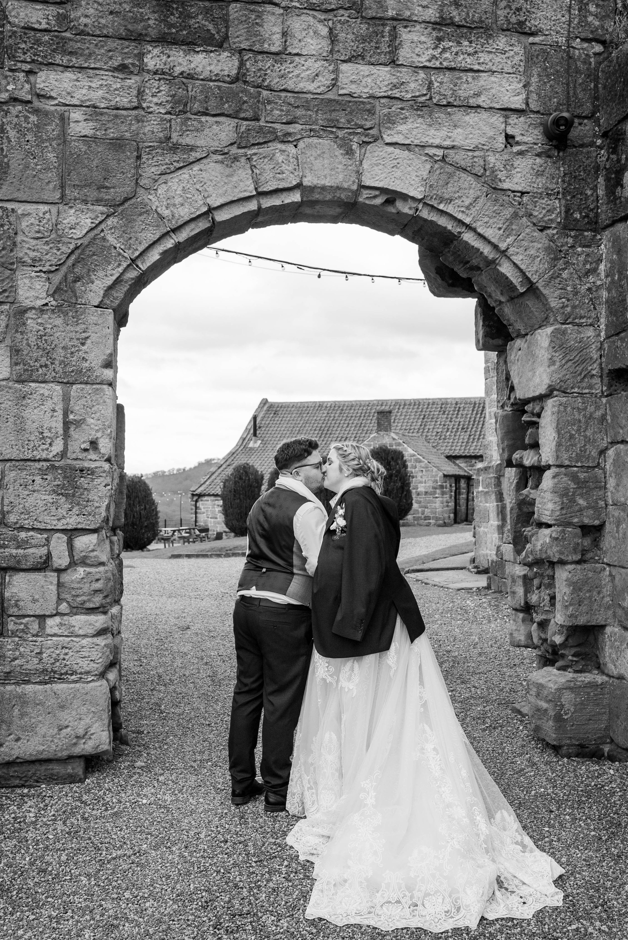 A black and white photo of a couple kissing under an arched stone gateway outdoors, with a rural landscape and a house in the background.