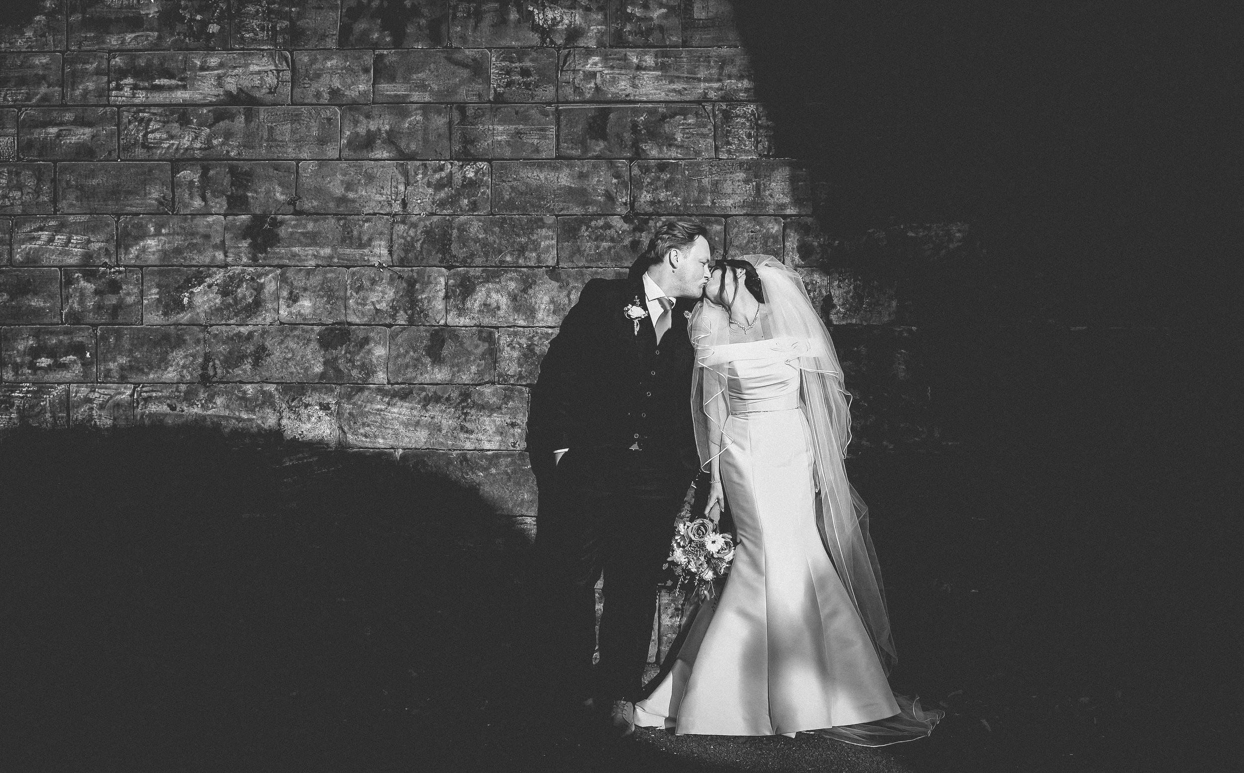 A black-and-white photo of a bride and groom, dressed in wedding attire, standing close together outdoors at night, with a brick wall in the background. The groom is holding a bouquet of flowers and they are sharing an intimate moment.