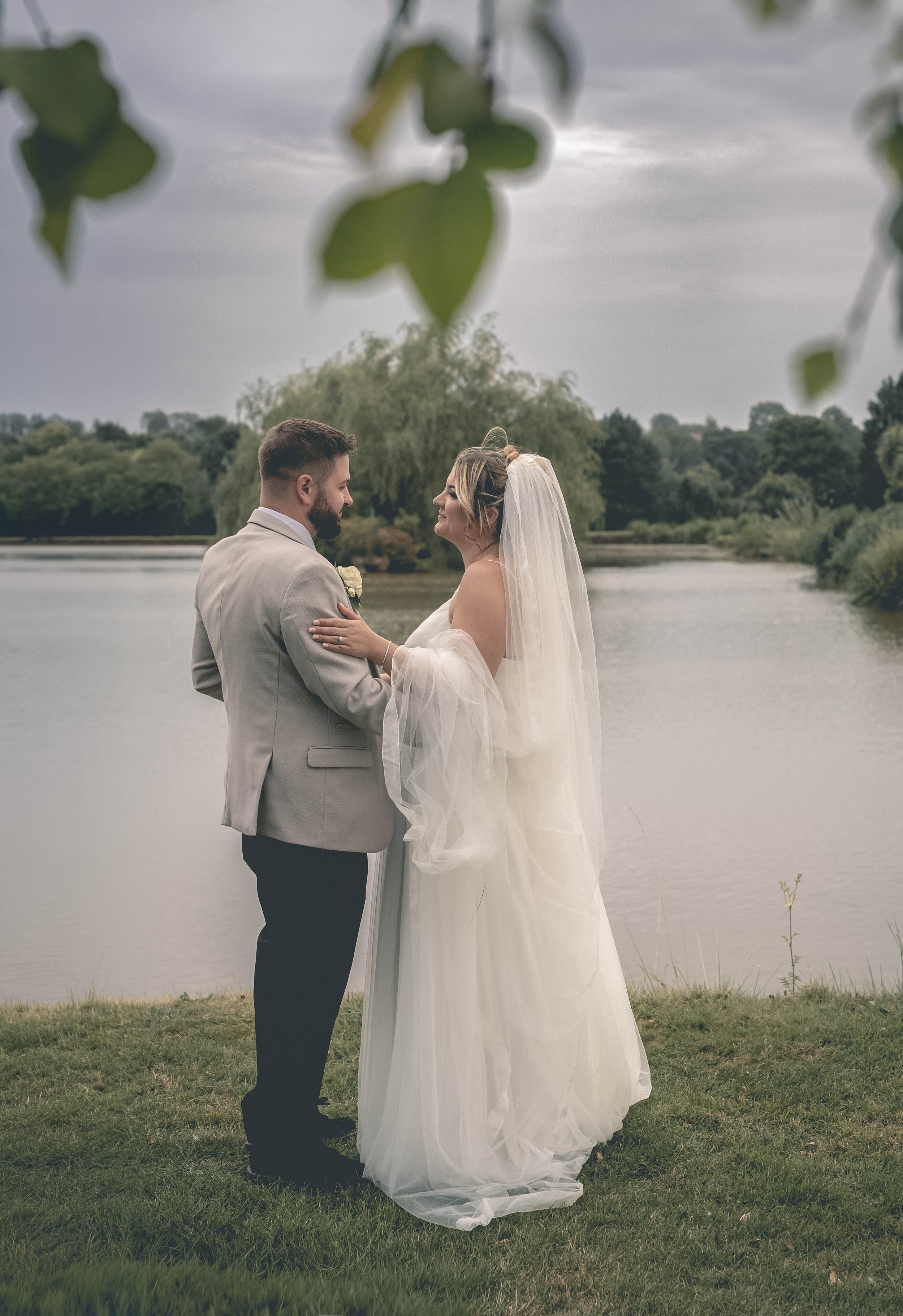 A bride and groom standing by a lake during their wedding, holding hands and gazing into each other's eyes, with overcast sky and lush greenery in the background.