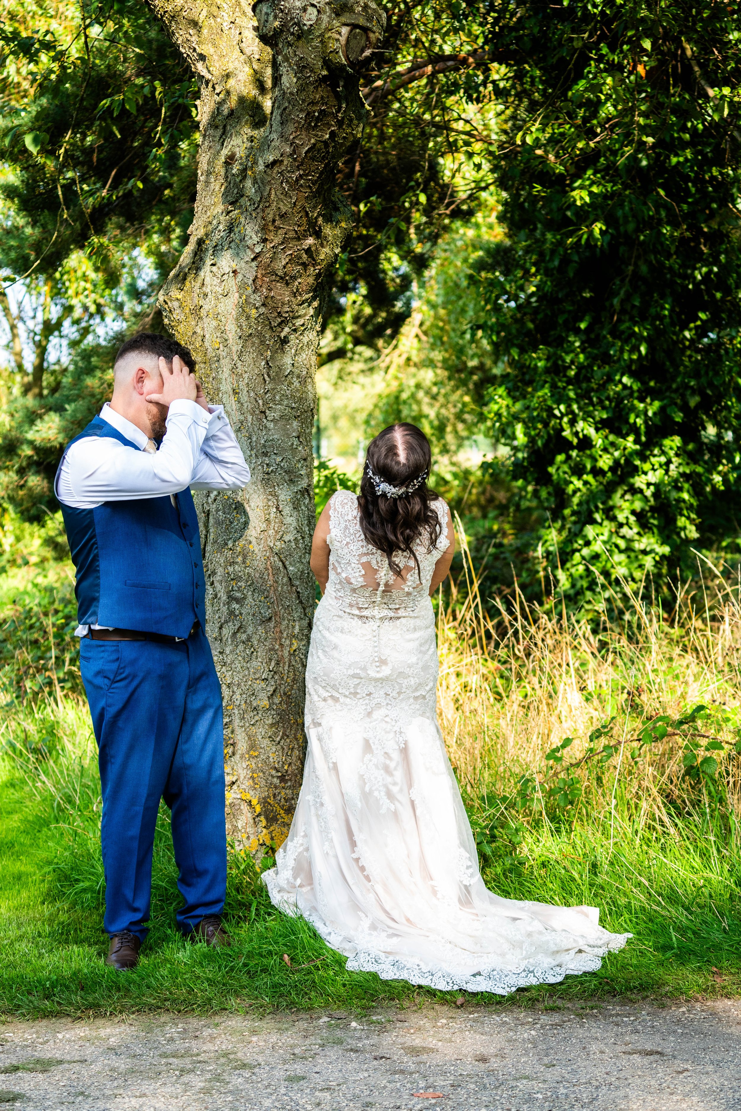 A bride in a white lace wedding dress stands with her back to a large tree, while a man in a blue suit, possibly the groom, stands nearby with his hands on his face, appearing distressed, in a lush outdoor setting.