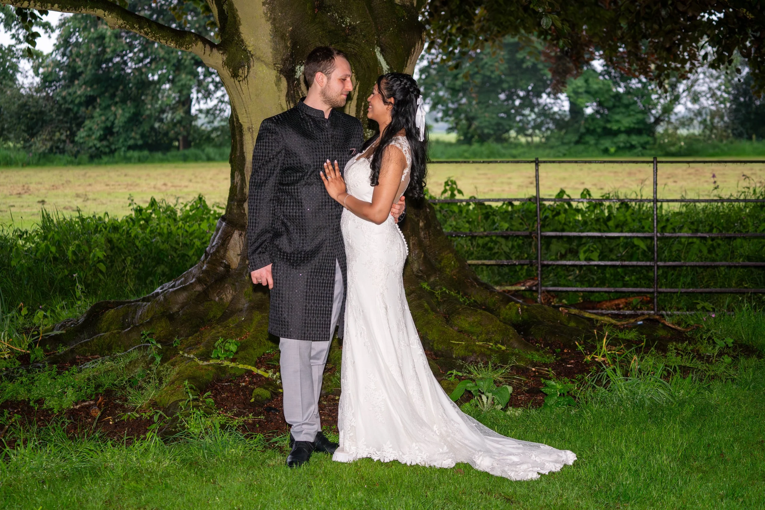 A bride wearng a white wedding dress and a groom in a black traditional coat, standing close by a large tree in a park, looking at each other.
