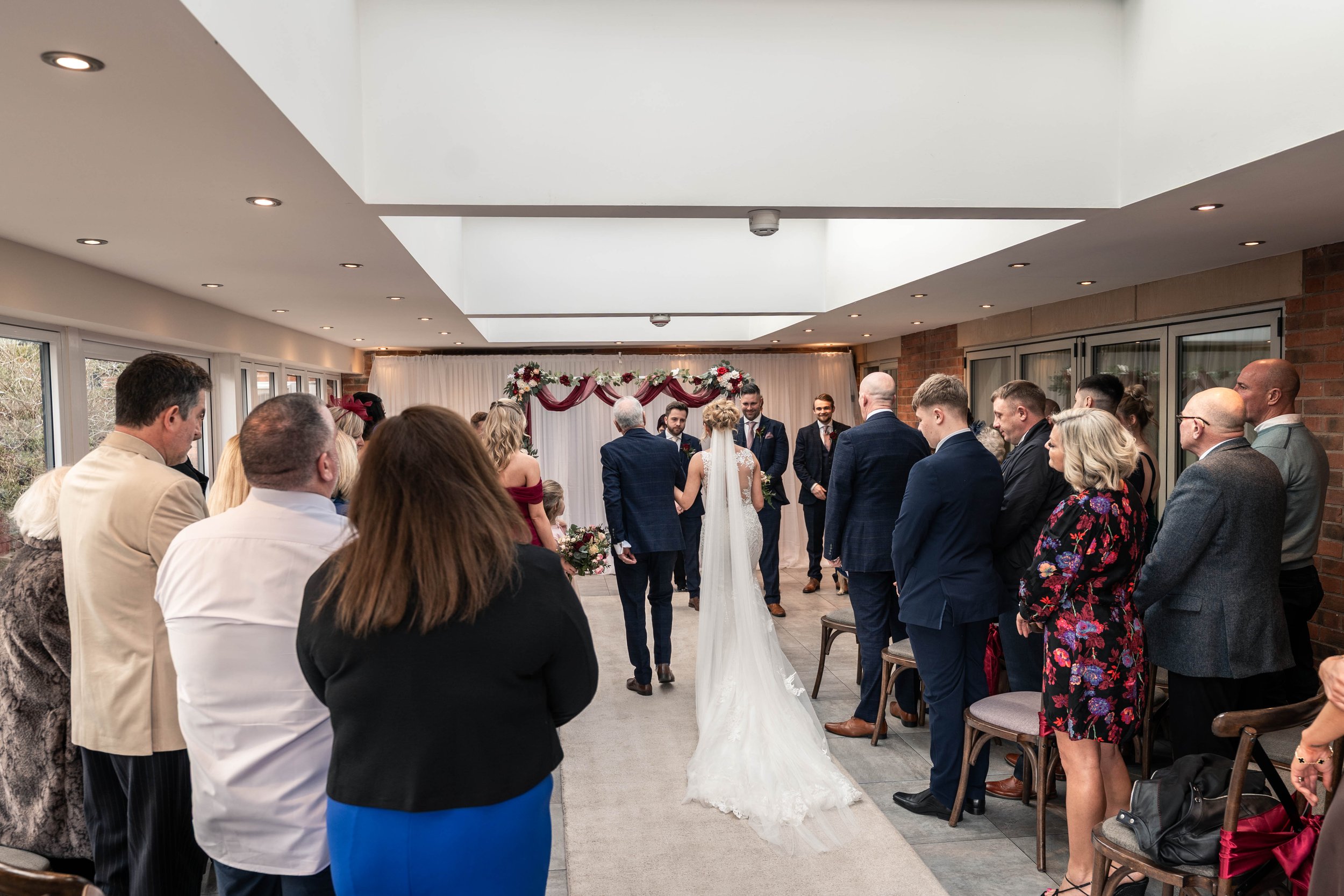 A wedding ceremony taking place indoors with the bride and groom standing at the altar, facing a priest or officiant, as family and friends stand around watching.