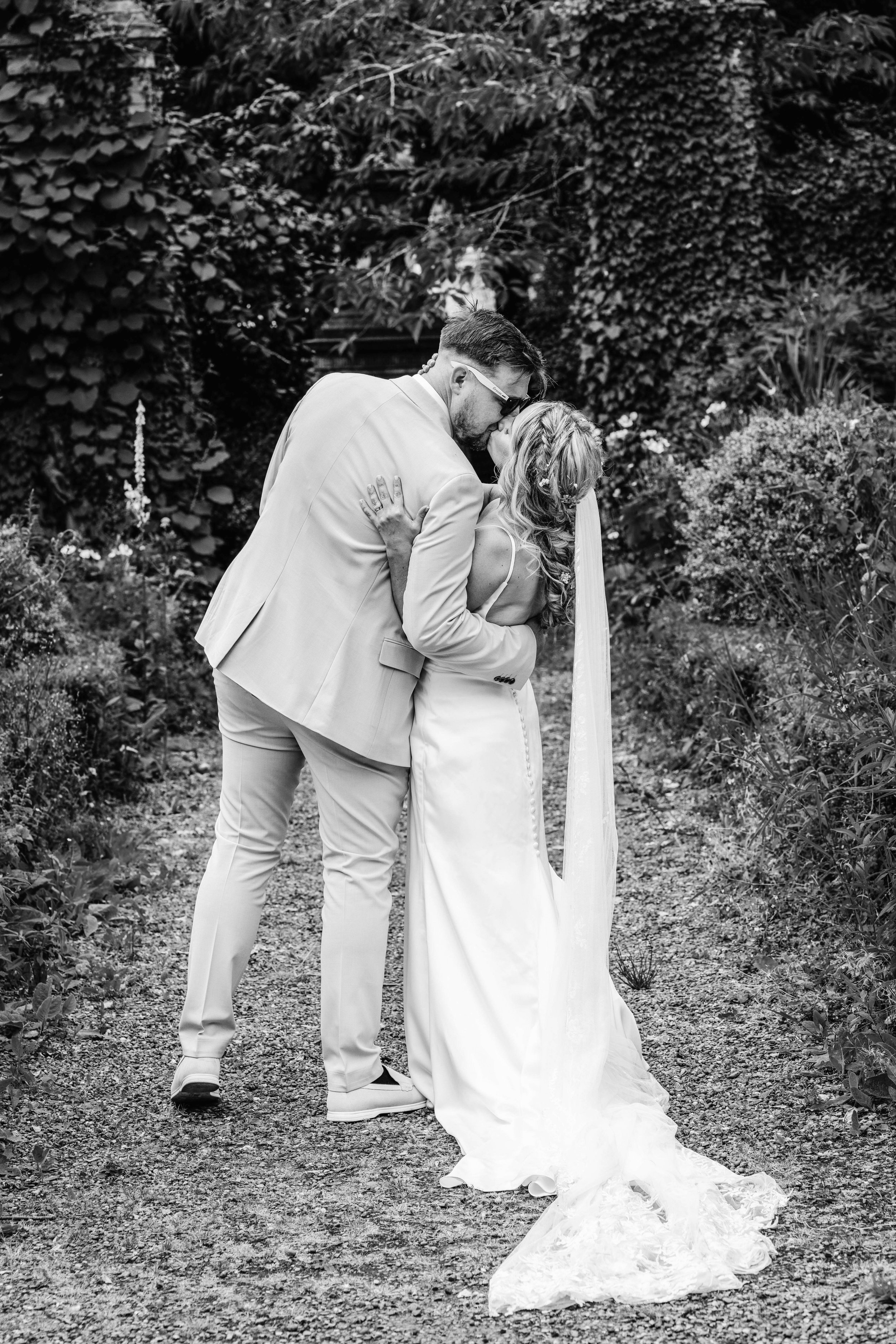 A black and white photo of a newly married couple sharing a kiss outdoors, with lush plants and trees in the background. The groom wears sunglasses and a suit, and the bride wears a flowing wedding gown and veil.