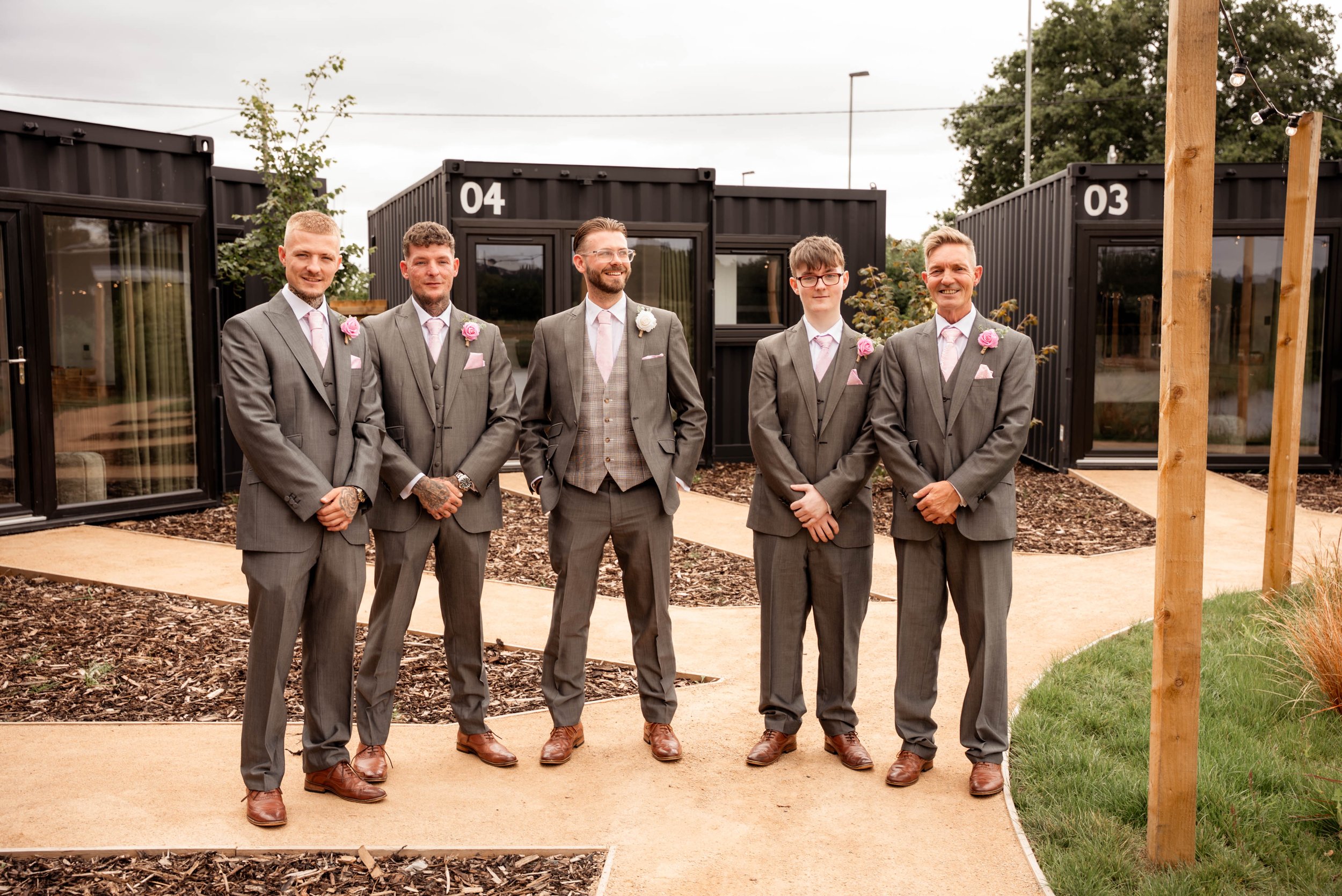 Six men in gray suits and pink ties standing outdoors on a curved walkway with black container-like buildings in the background, smiling and posing for a photo.