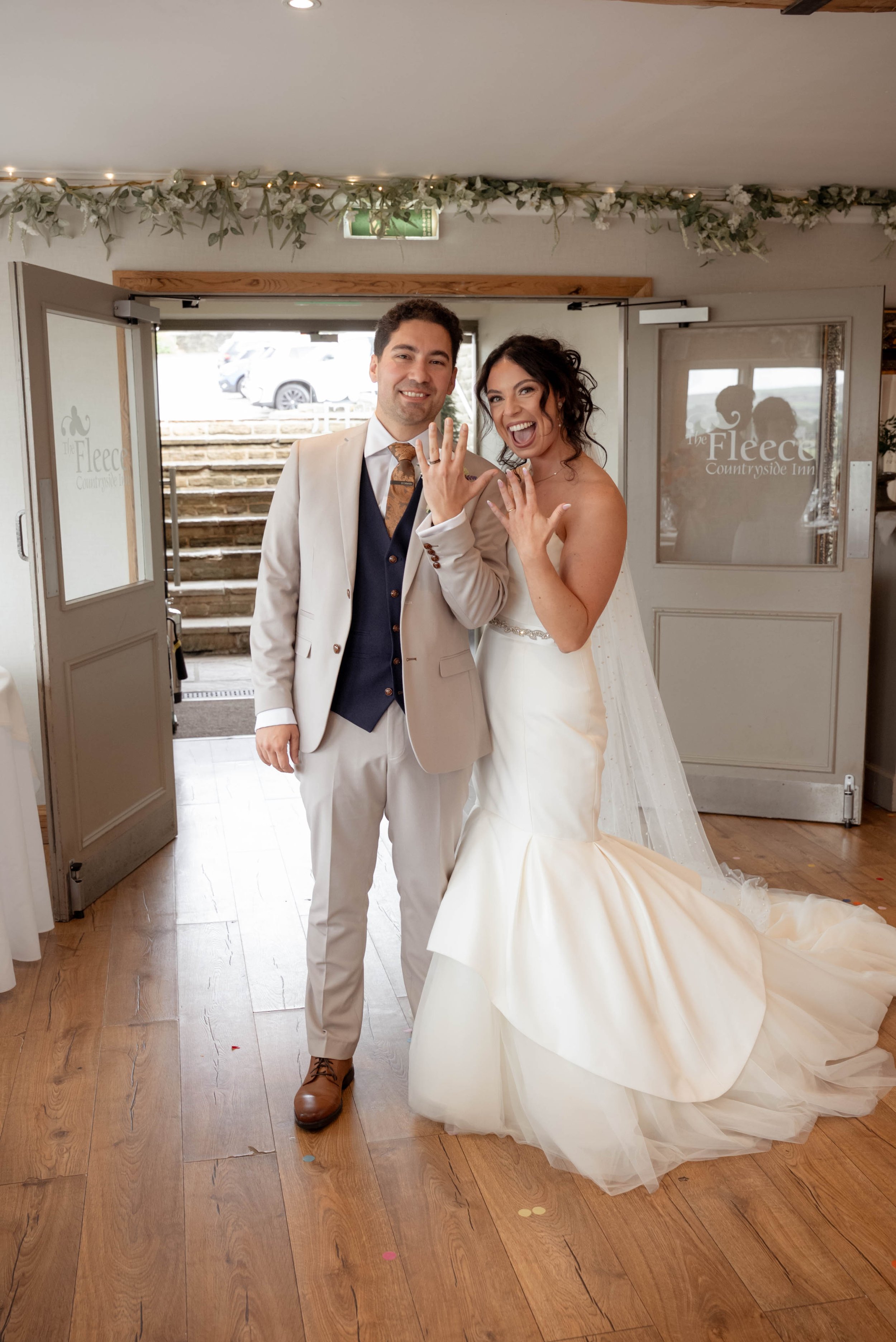 A newlywed couple showing their wedding rings, smiling Joyfully inside a rustic venue with wooden floors and greenery decorations.