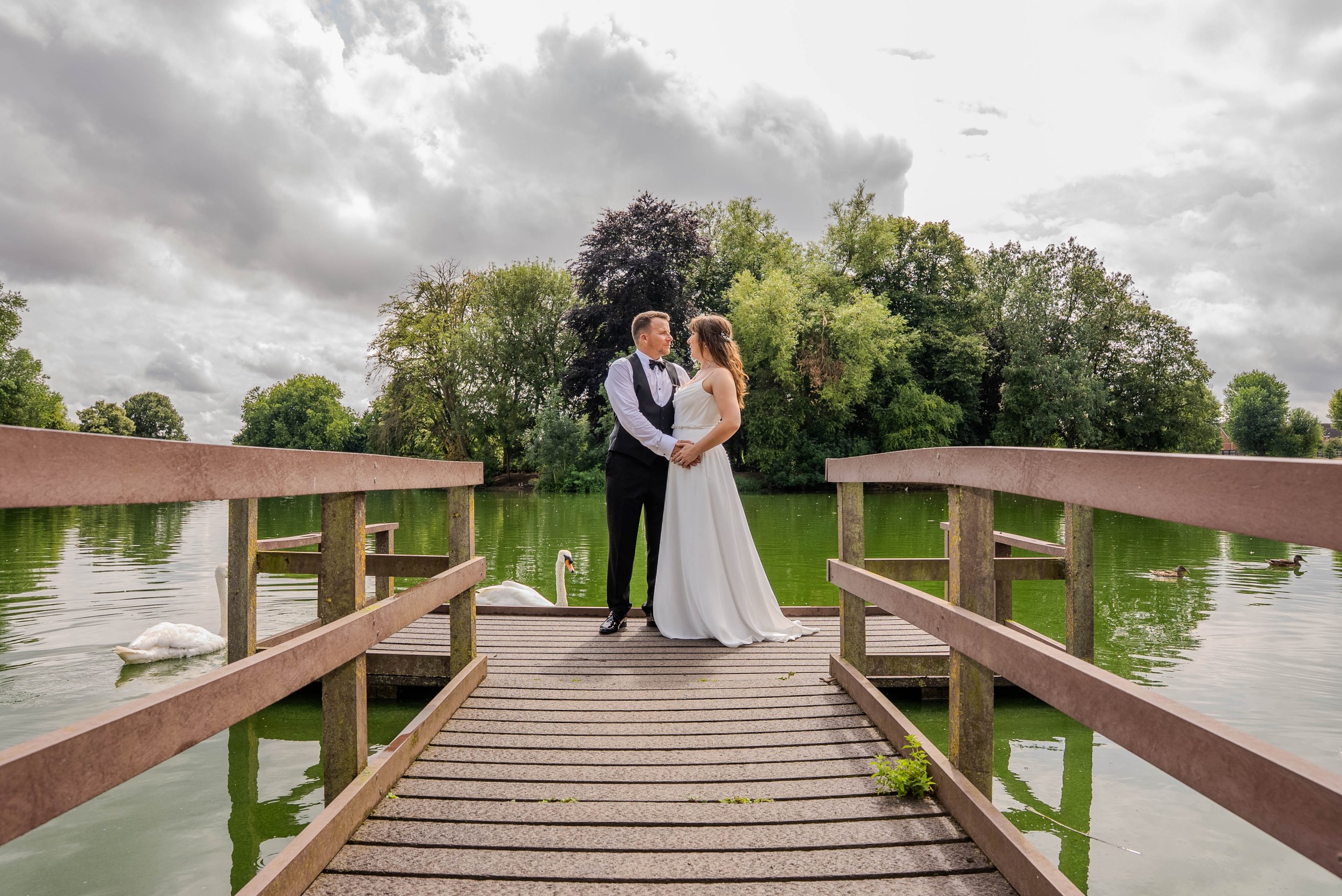 A bride and groom standing on a wooden dock by a lake, holding hands and gazing at each other, with swans swimming nearby and trees in the background under a cloudy sky.
