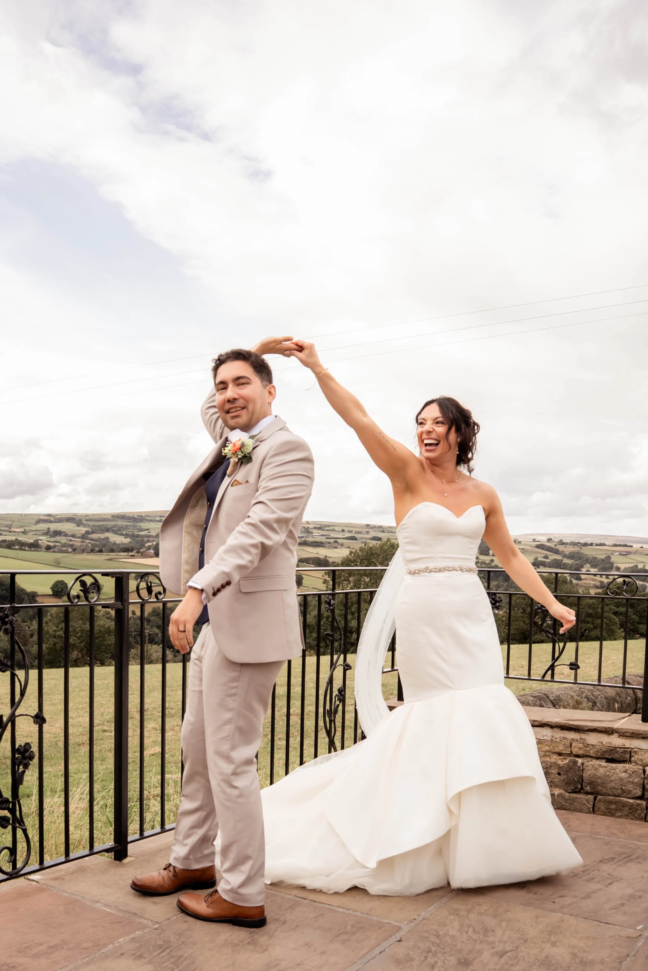 A bride and groom in wedding attire outdoors on a balcony with a scenic rural landscape in the background. The bride is playfully holding a hat or a piece of fabric above the groom's head, and both are smiling.