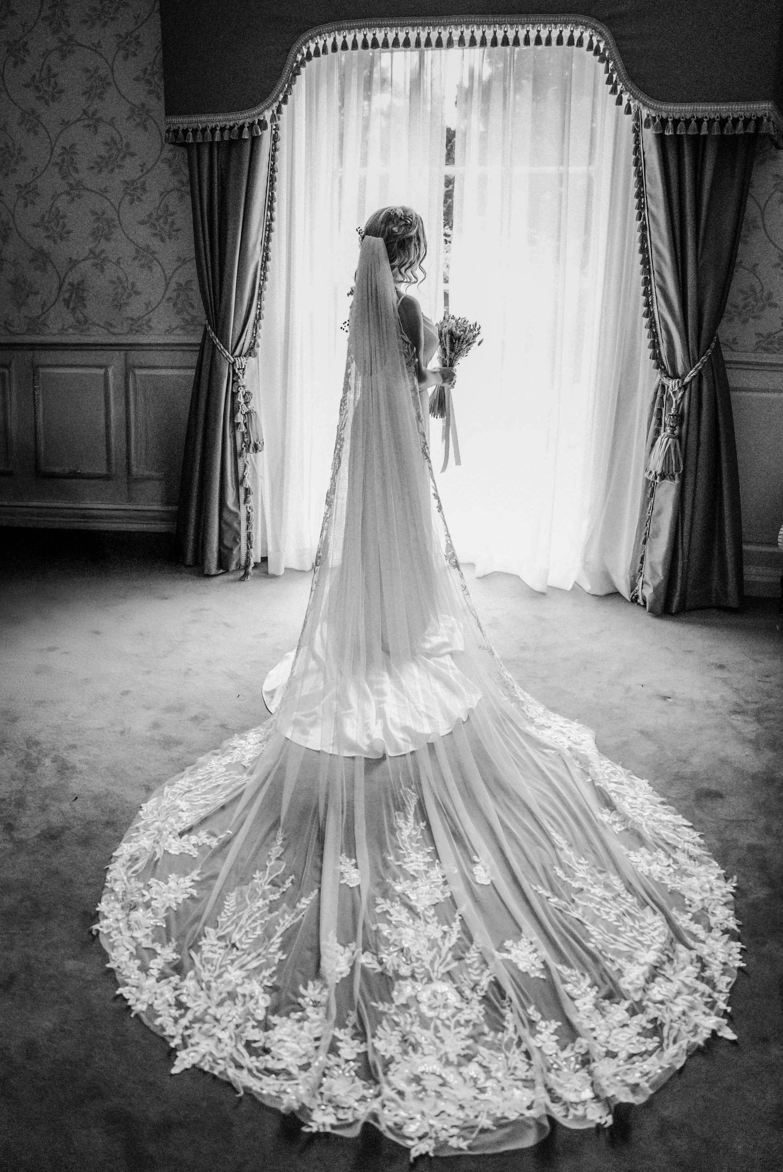 A bride in a wedding gown with a long, embroidered train stands near a window, holding a bouquet, in a room with curtains and patterned wallpaper.