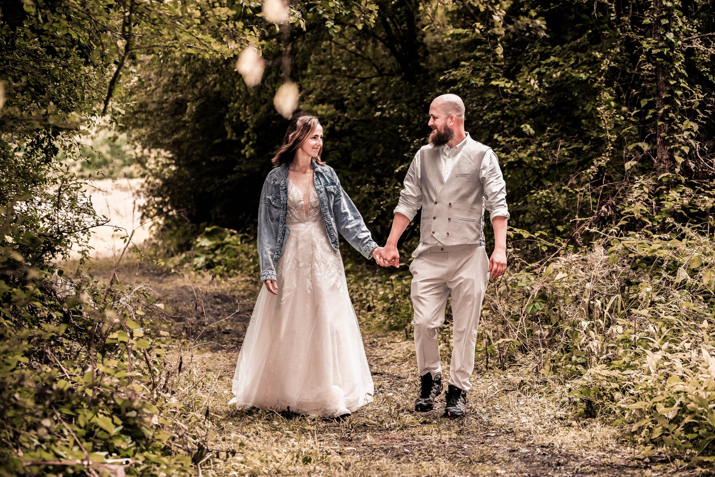 A couple walking hand in hand on a trail in a wooded area, dressed in wedding attire, smiling at each other.