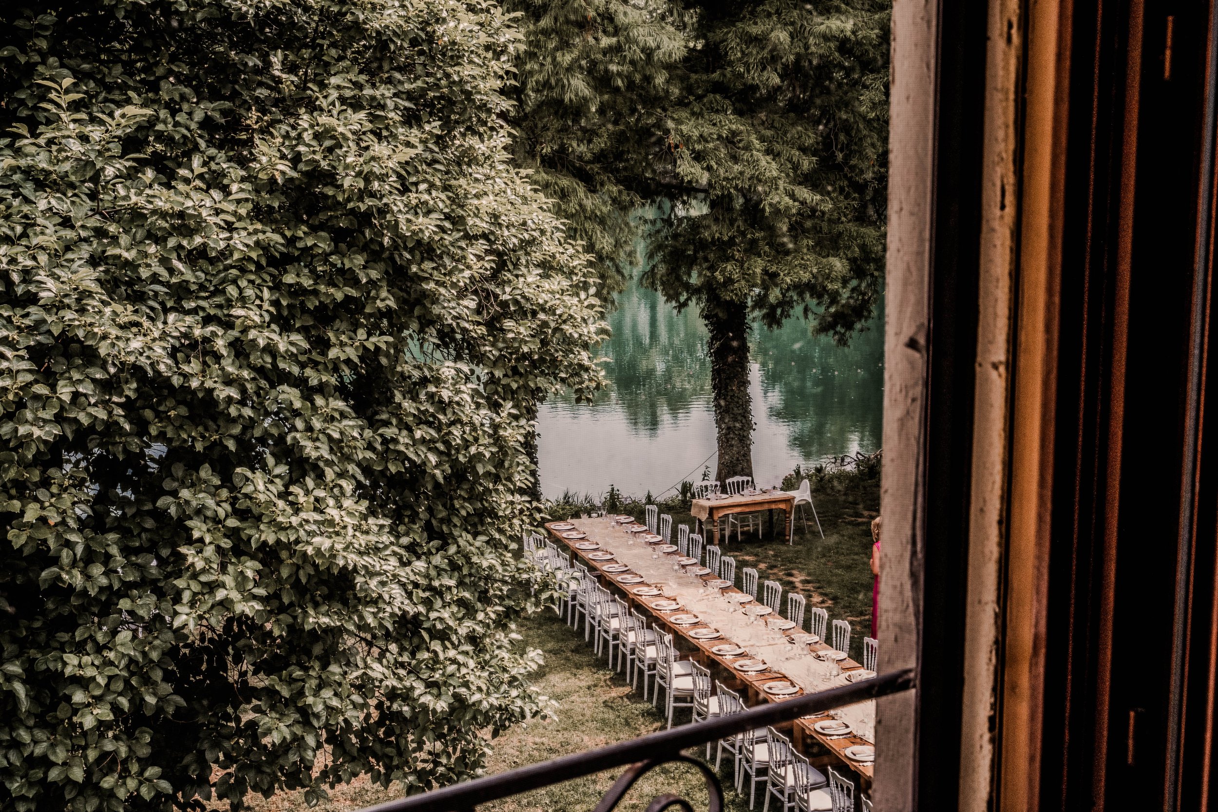 Long dining table set outdoors by a lake, viewed from a window, with trees surrounding and additional smaller tables in the background.