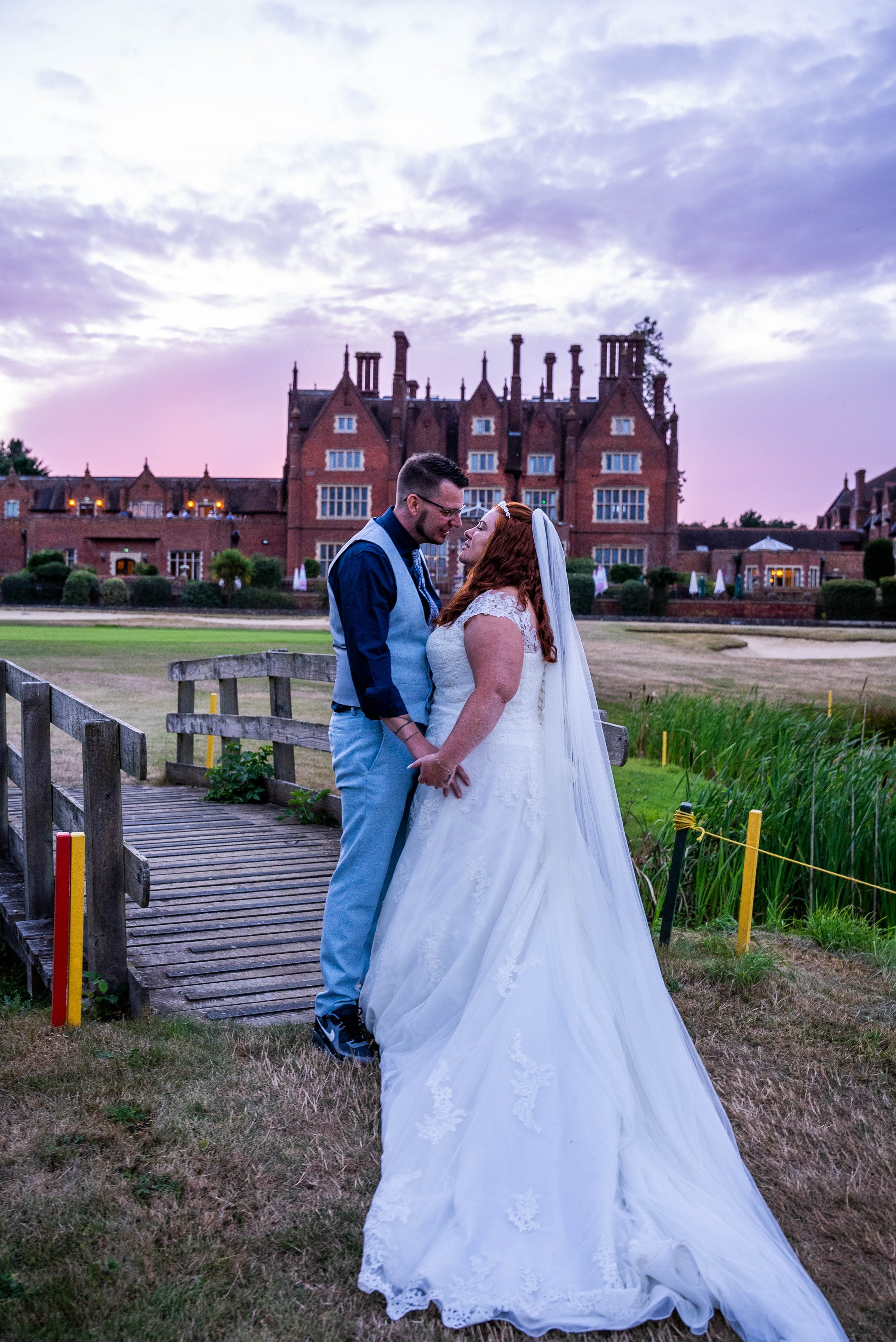 A bride and groom stand close together on a small wooden bridge outdoors, with a large historic brick building and a partly cloudy sky at sunset in the background.