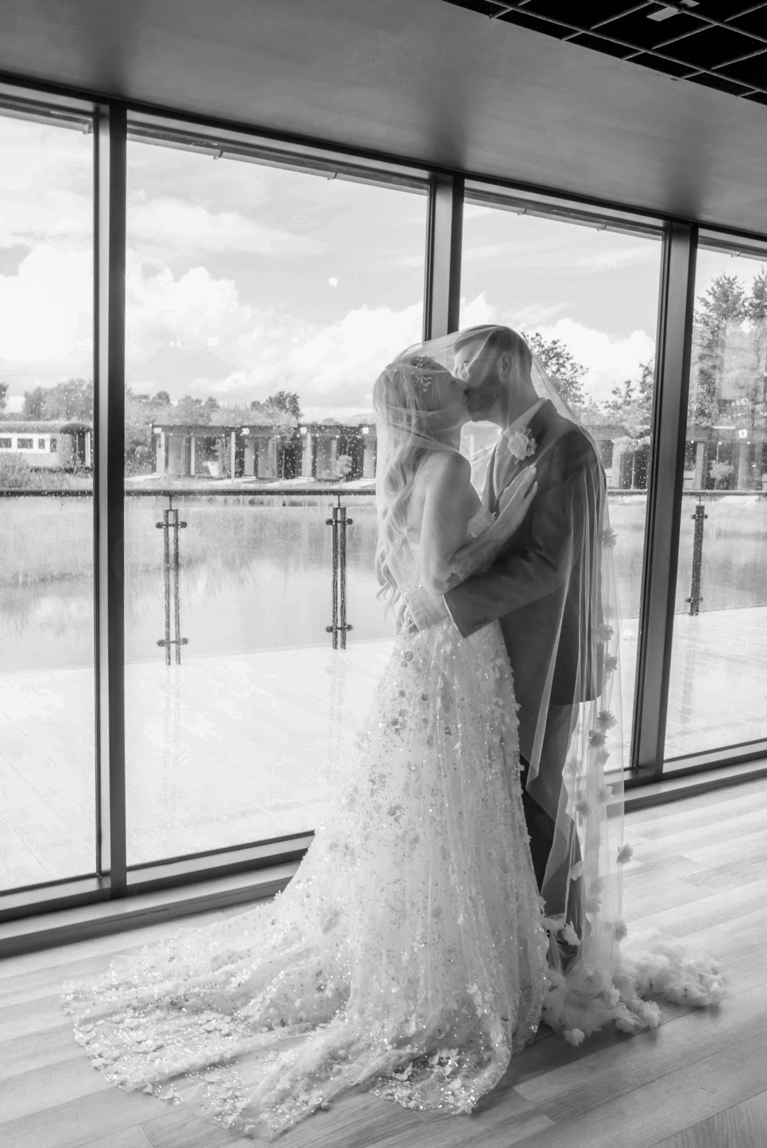 Black-and-white photo of a bride and groom kissing behind a glass window, with a rainy outdoor view in the background.