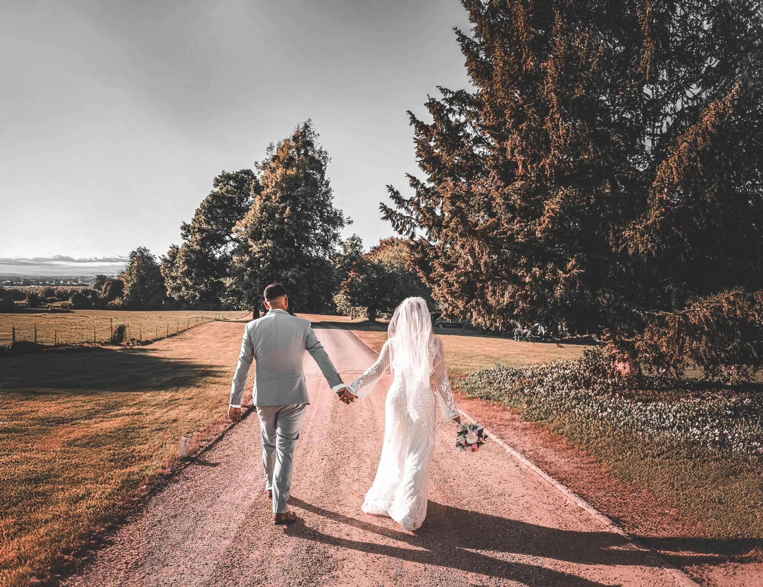 A bride and groom walk hand in hand on a dirt road in a scenic rural area, with large trees on the right and rolling fields in the background.