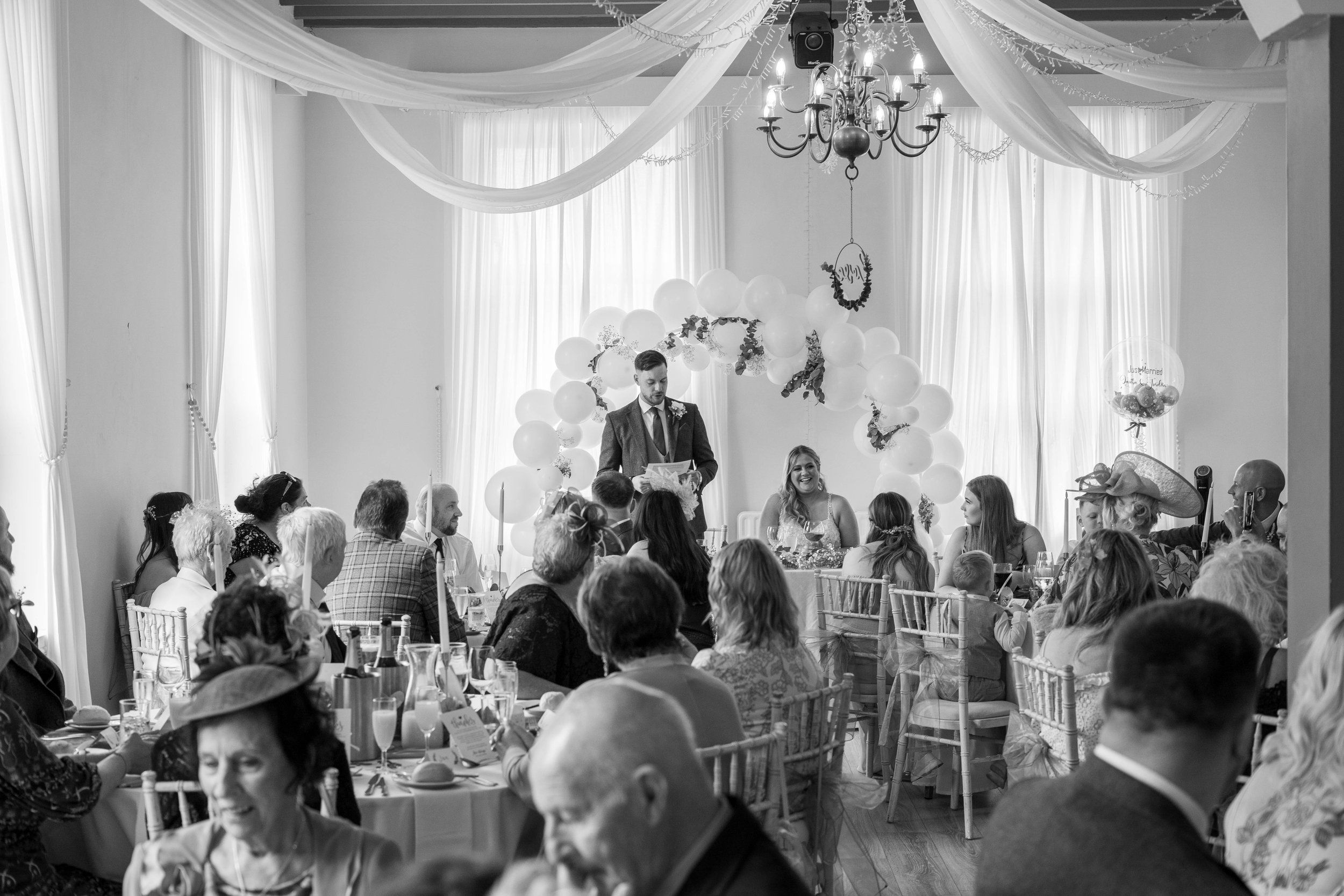 A black-and-white photo of a wedding reception with guests seated at tables, a bride and groom sitting at a table, and a man in a suit giving a speech at the front of the room. The room is decorated with draped fabric, balloons, and a chandelier.