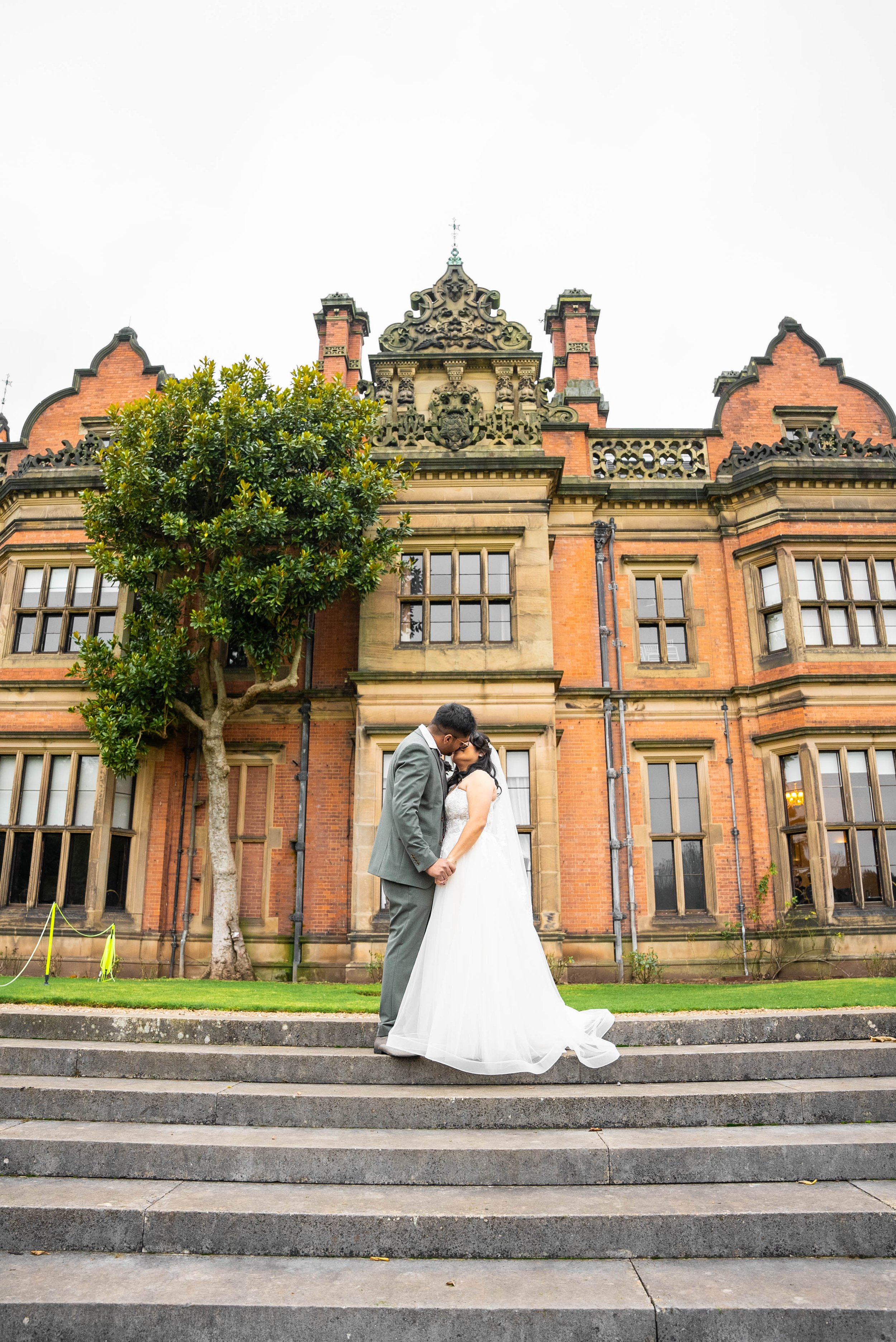 A bride and groom kissing and holding hands on a staircase outside a historic brick building with large windows and ornate architectural details, and a tree to the left.