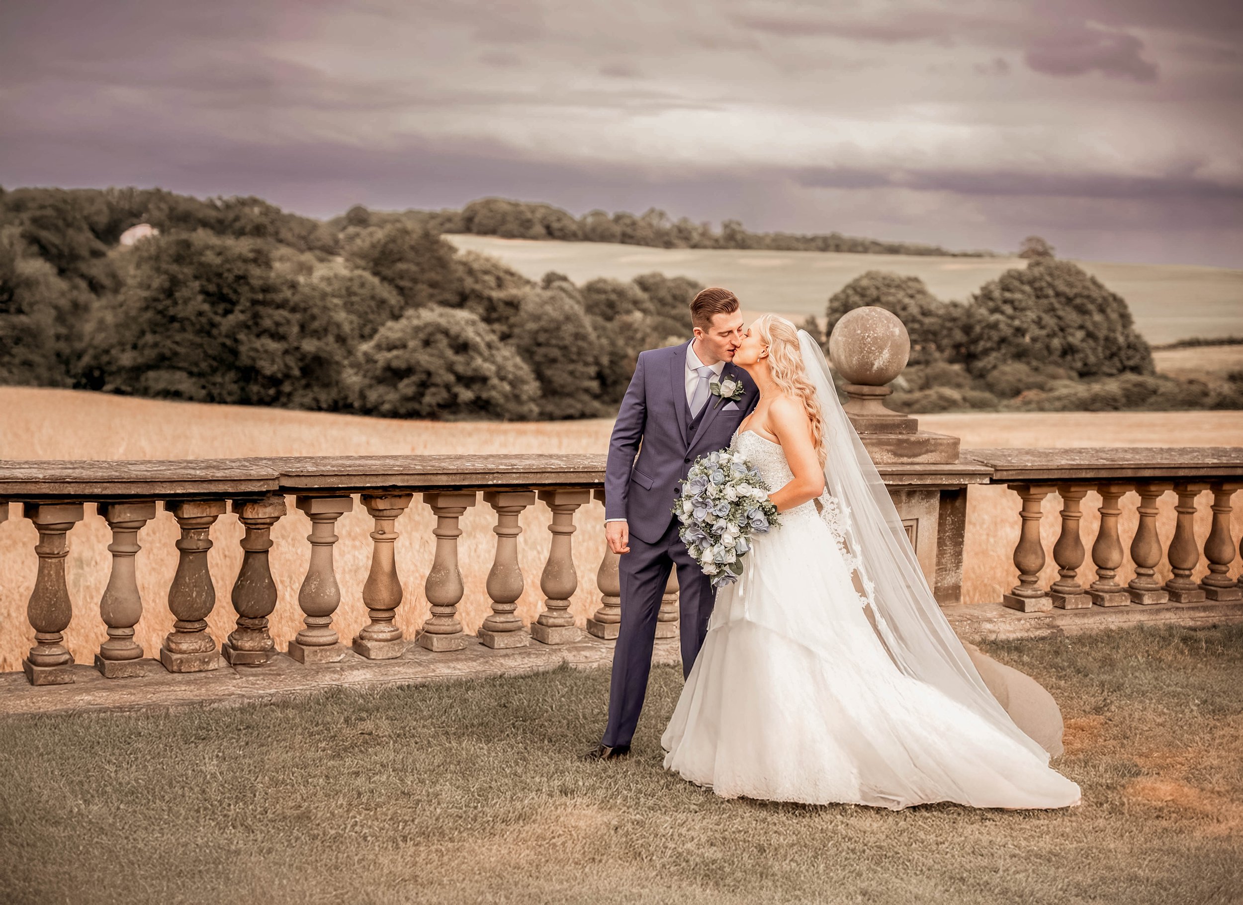A bride and groom sharing a kiss on a stone balcony overlooking a countryside landscape with rolling hills and trees during cloudy weather.