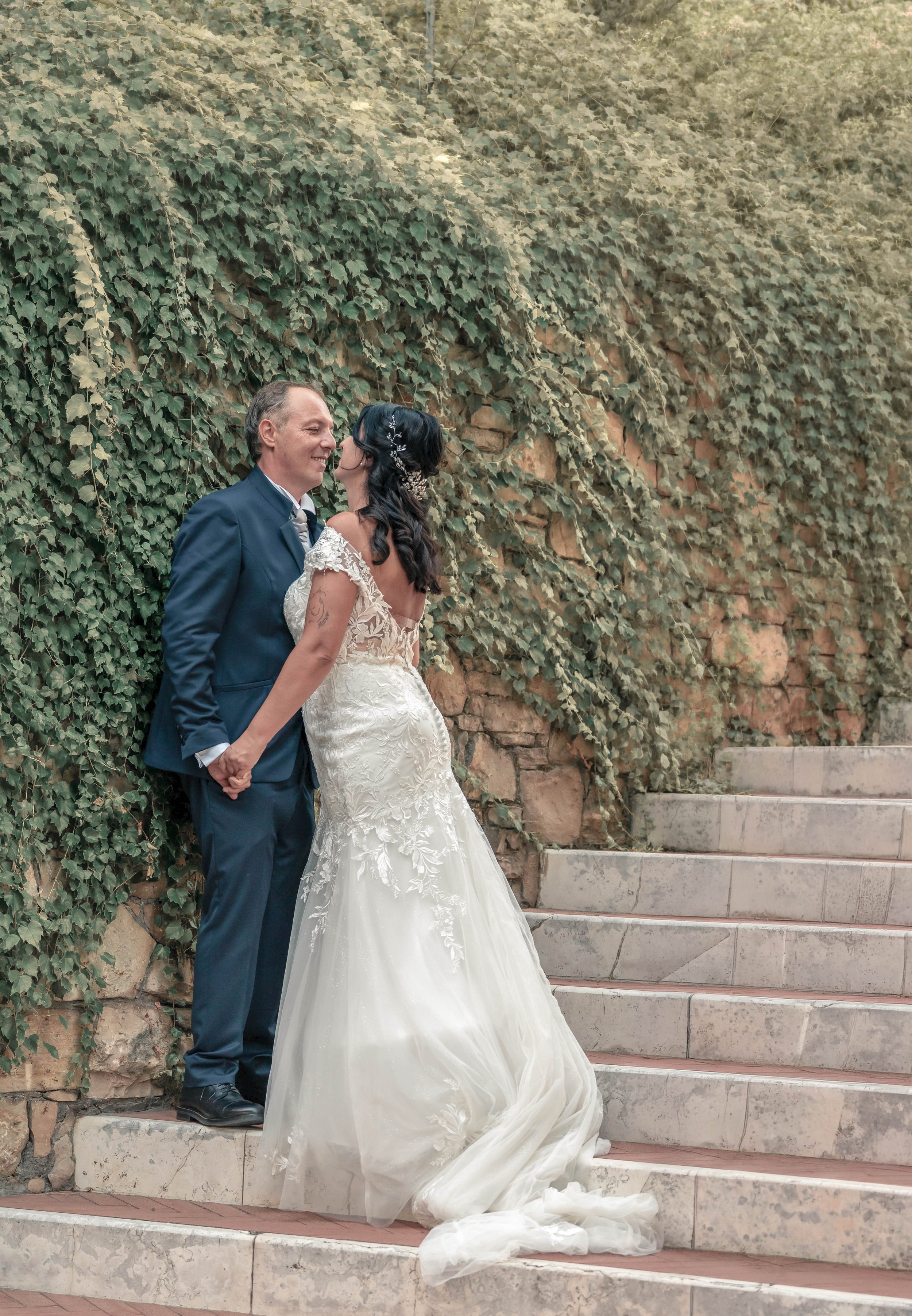 A bride and groom in wedding attire standing on outdoor stone steps with ivy-covered wall behind them. The bride wears a white lace wedding dress and the groom wears a navy blue suit. They are holding hands and leaning in for a kiss.