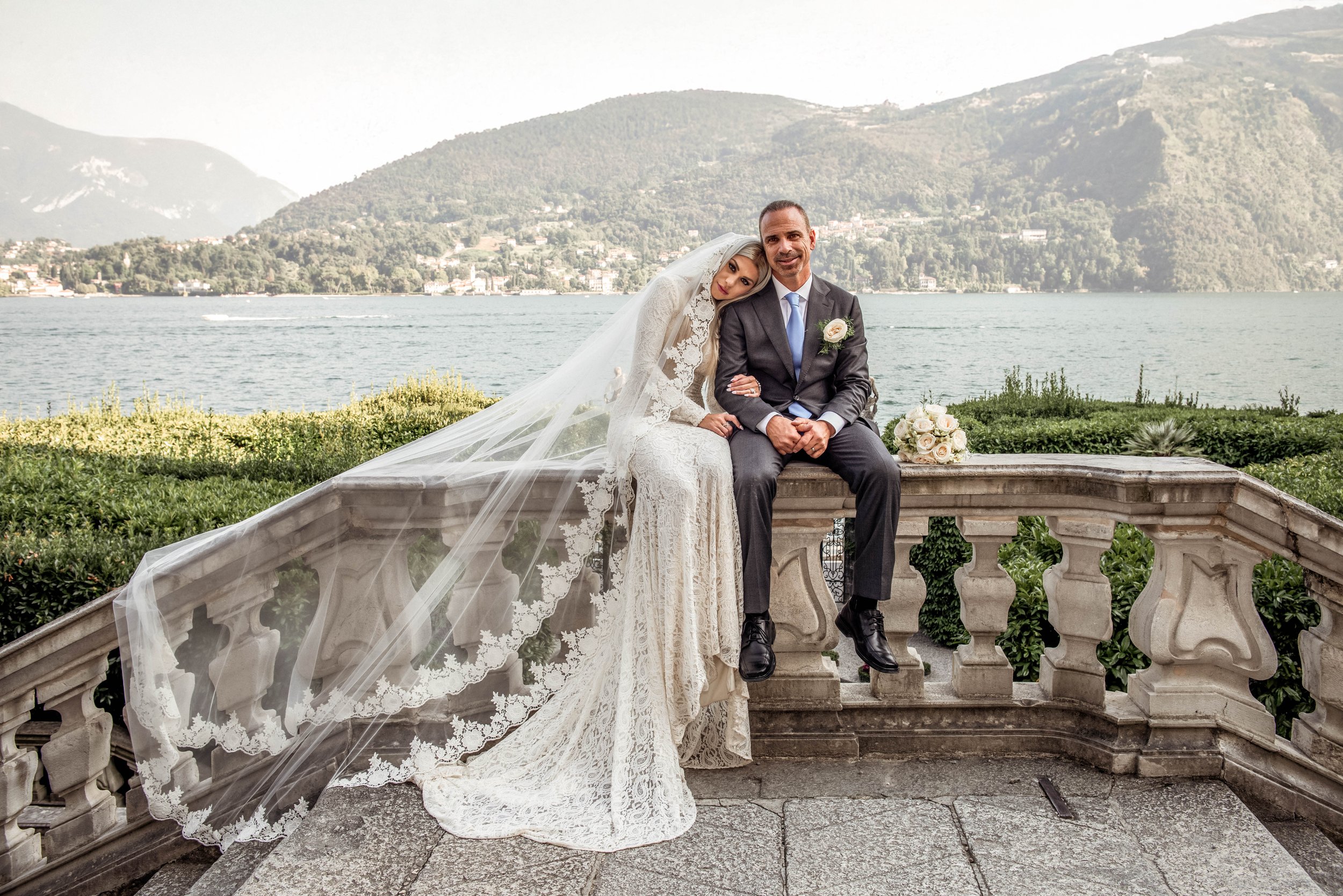 Bride and groom sitting on a stone balustrade outdoors with lake, mountains, and greenery in background, during wedding.