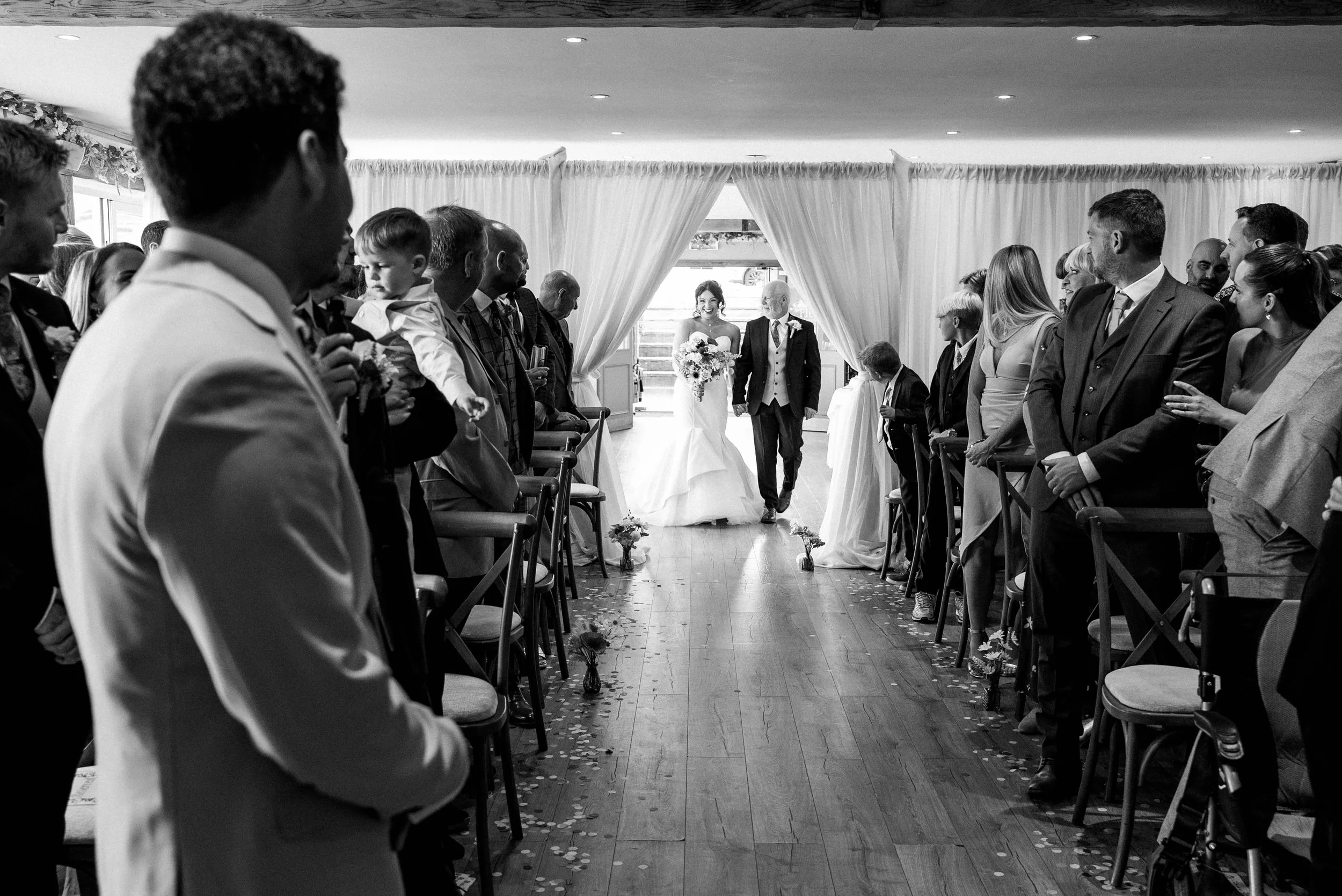 A bride and groom walking down the aisle at their wedding ceremony, surrounded by seated guests on either side, with some guests standing. The venue has large curtains, and flower petals are scattered on the floor.