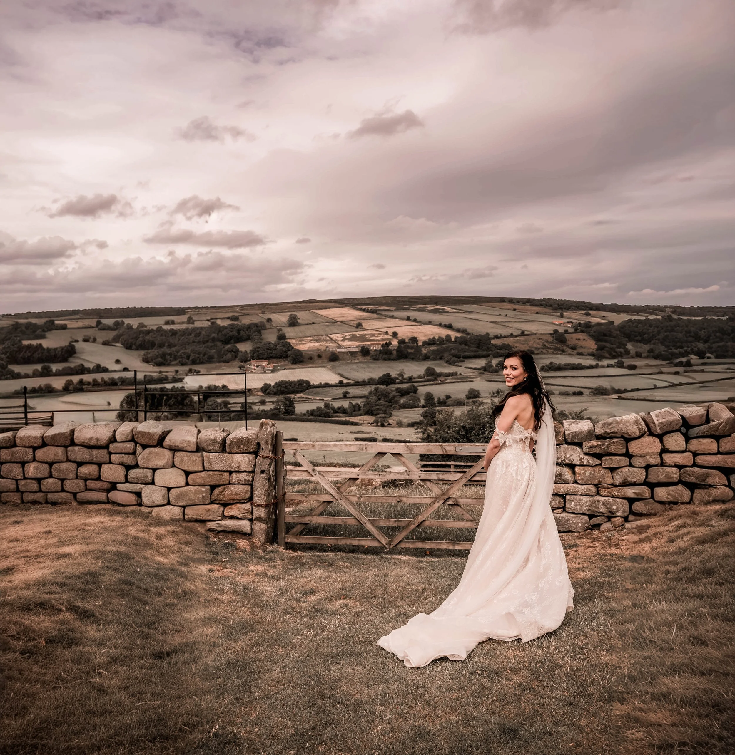 A bride in a wedding dress standing on a grassy area in front of a stone wall and wooden gate, with rolling countryside and cloudy sky in the background.