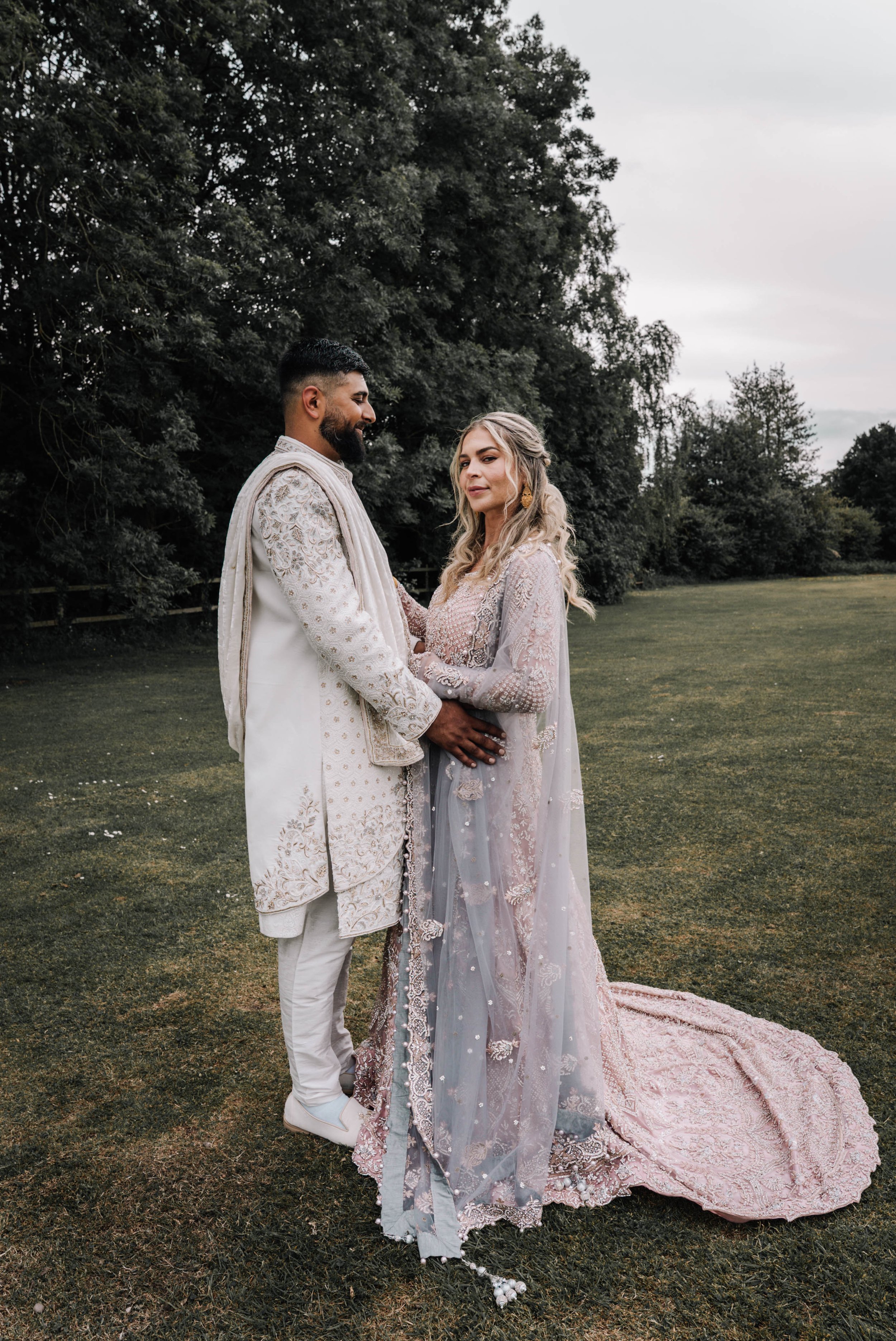 A bride and groom holding hands outdoors in a grassy area, with trees in the background. The groom is wearing a cream-colored traditional outfit with embroidery, and the bride is in a detailed, pastel-colored wedding dress with lace and embellishment