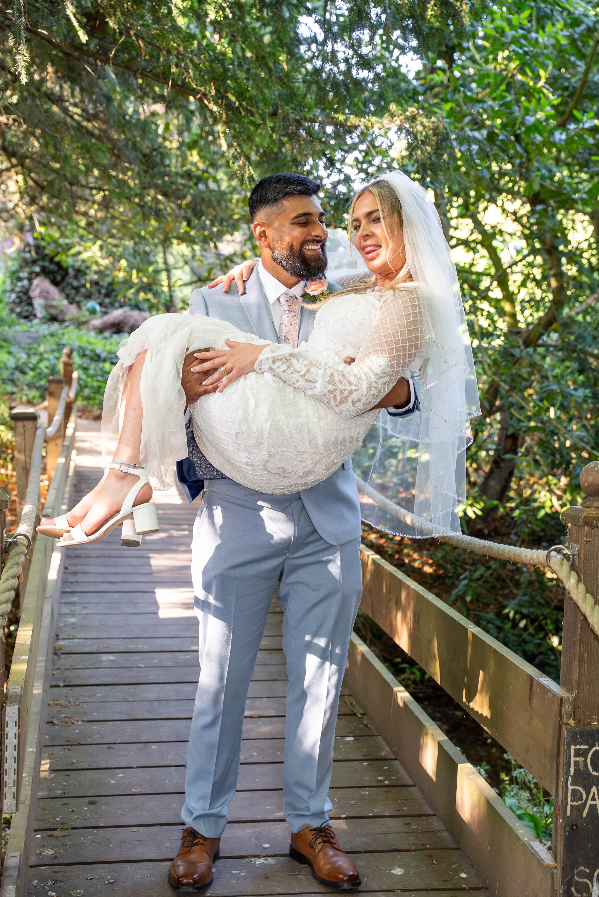 A man in a light gray suit and a woman in a white wedding dress and veil enjoying a moment together on a wooden bridge in a lush green forest. The man is holding the woman in his arms.