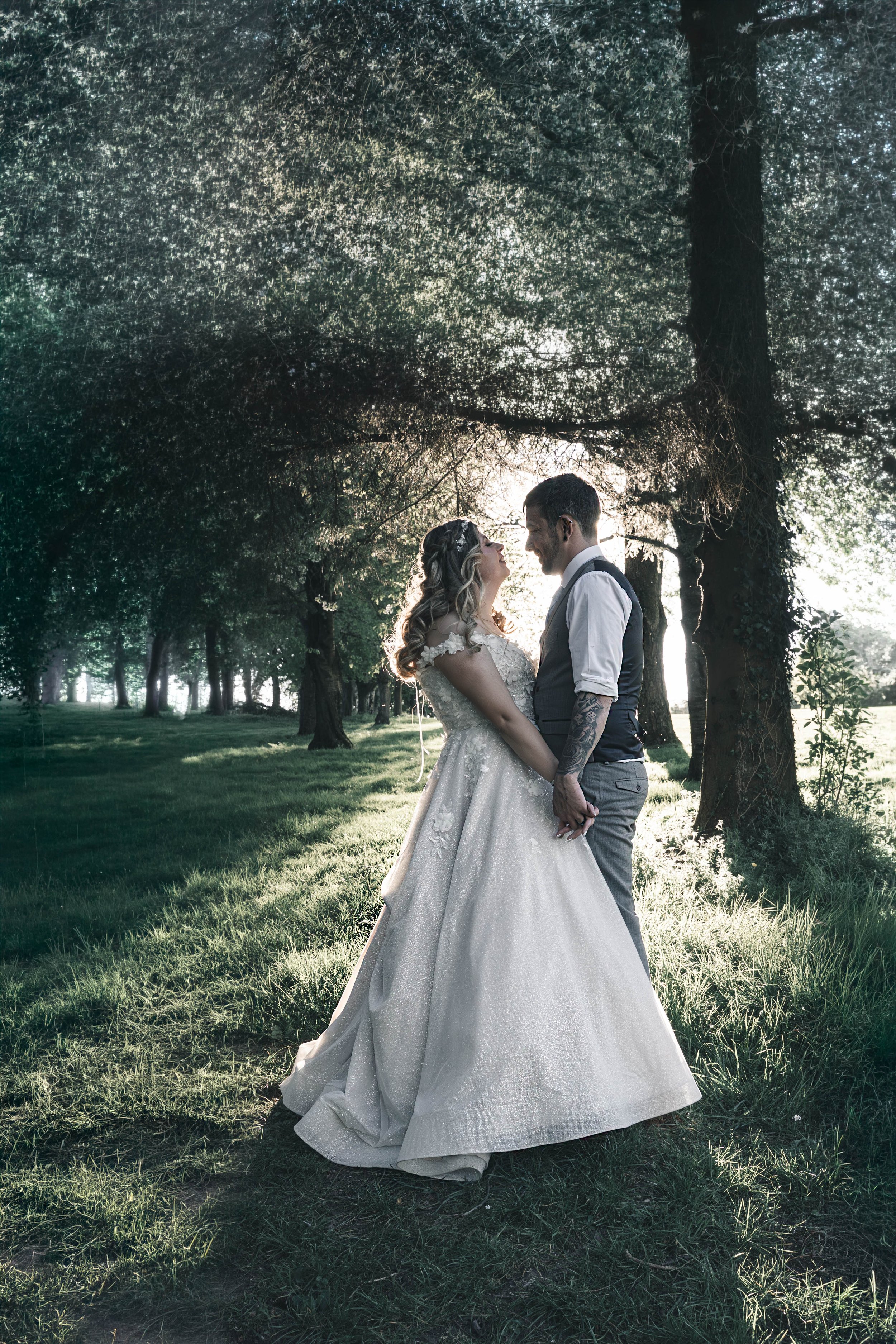 A bride and groom standing close and smiling at each other outdoors in a wooded park during sunset. The bride is wearing a white lace wedding dress, and the groom is dressed in a vest and trousers. They are holding hands with sunlight shining behind 