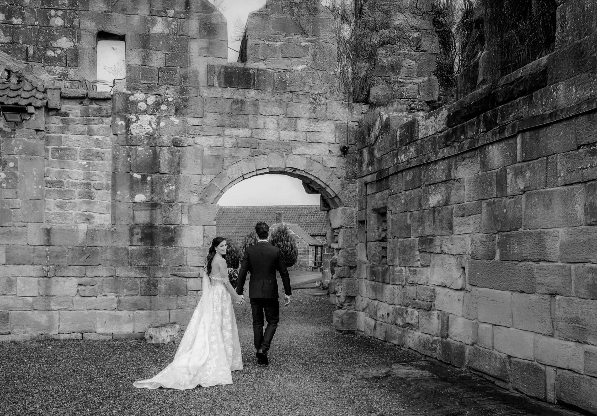 Black and white photo of a bride and groom walking hand in hand through a stone archway, with the bride smiling at the groom. The bride is wearing a long lace wedding gown, and the groom is in a dark suit. The scene appears to be outdoors near old st