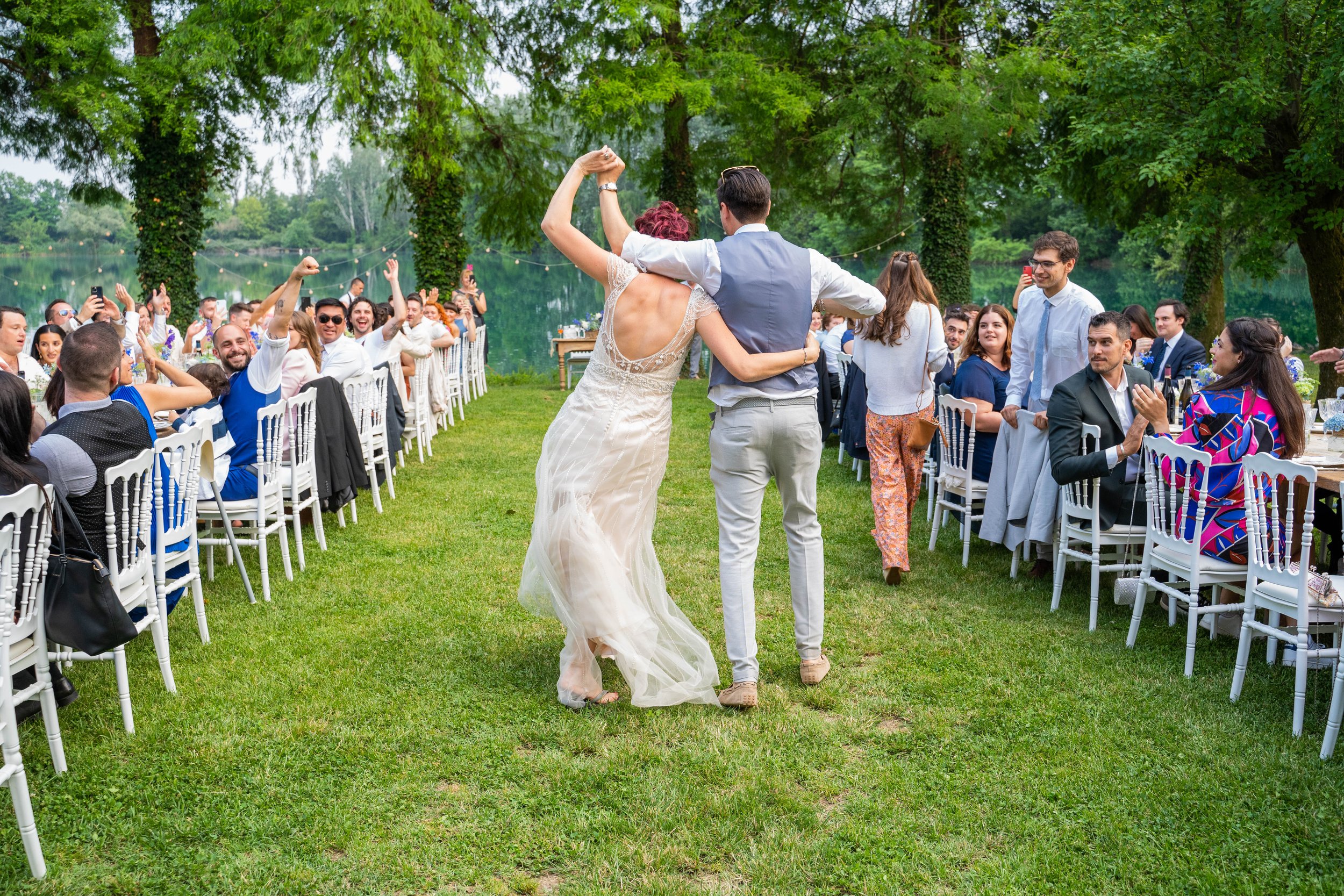 People attending an outdoor wedding reception by a lake, with a couple dancing in the center and guests sitting at decorated tables, surrounded by large trees.