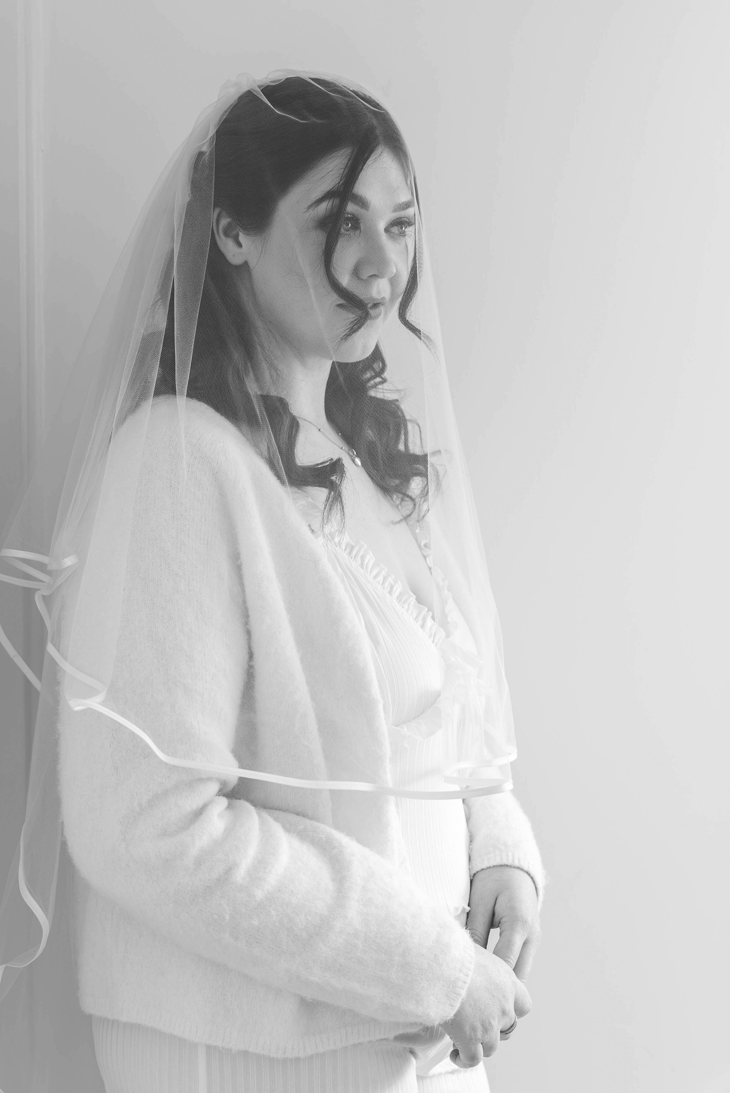 A woman wearing a wedding veil and white sweater, looking to the side with a calm expression, against a plain wall.