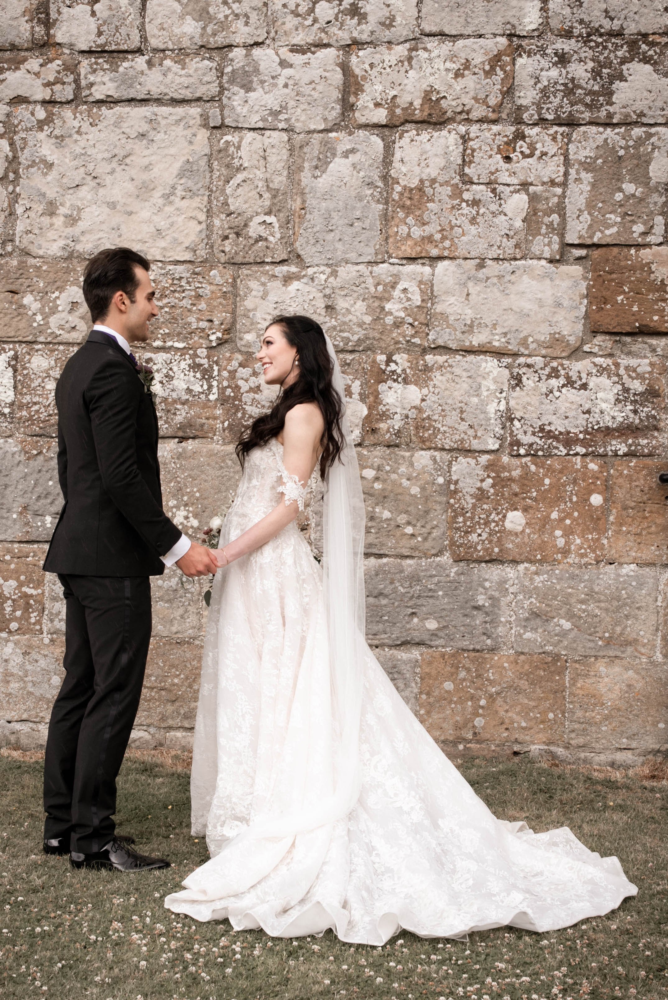 A bride and groom holding hands and smiling at each other outdoors against a stone wall.