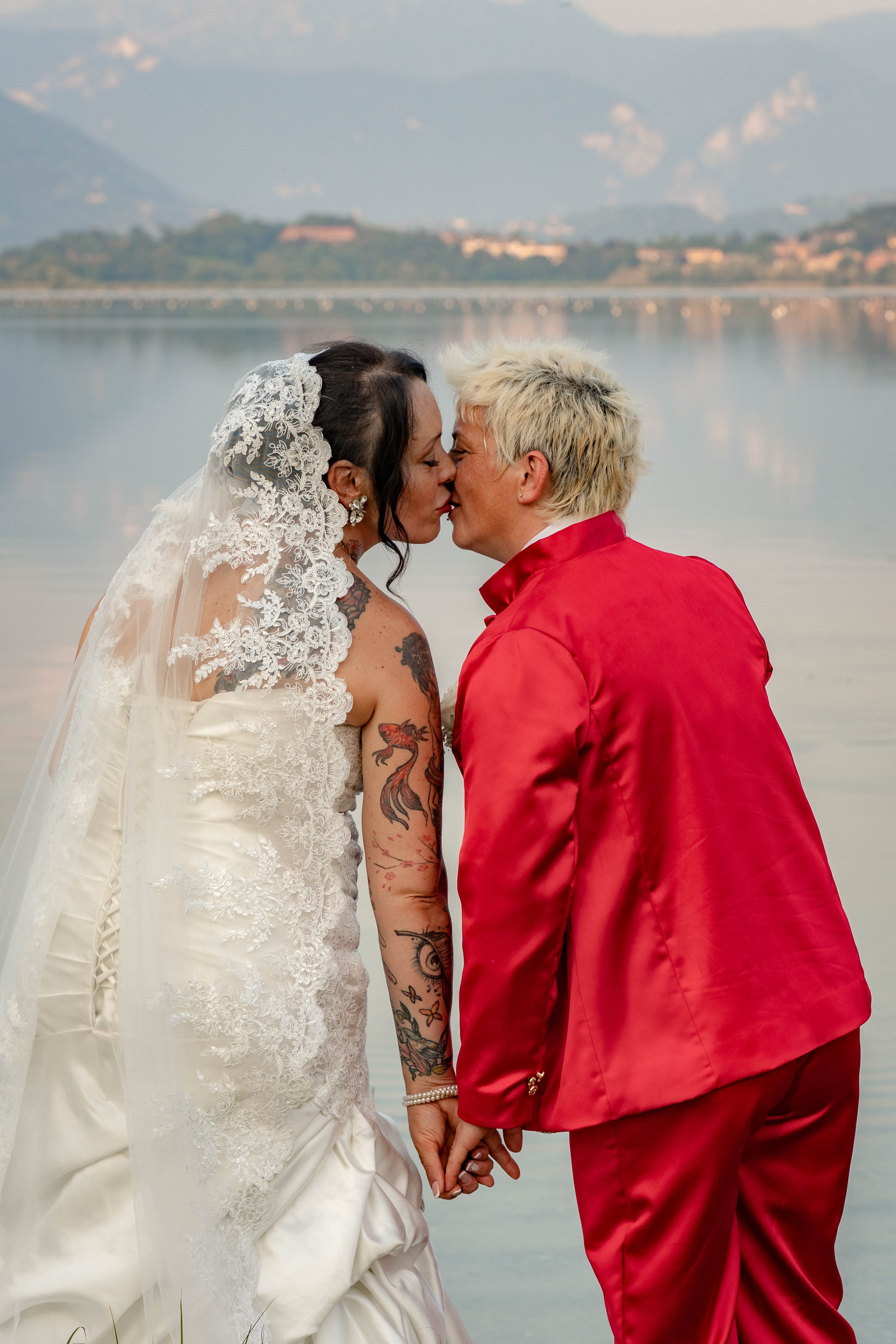 A couple dressed in wedding attire by a lake, about to kiss, holding hands, with mountains in the background.