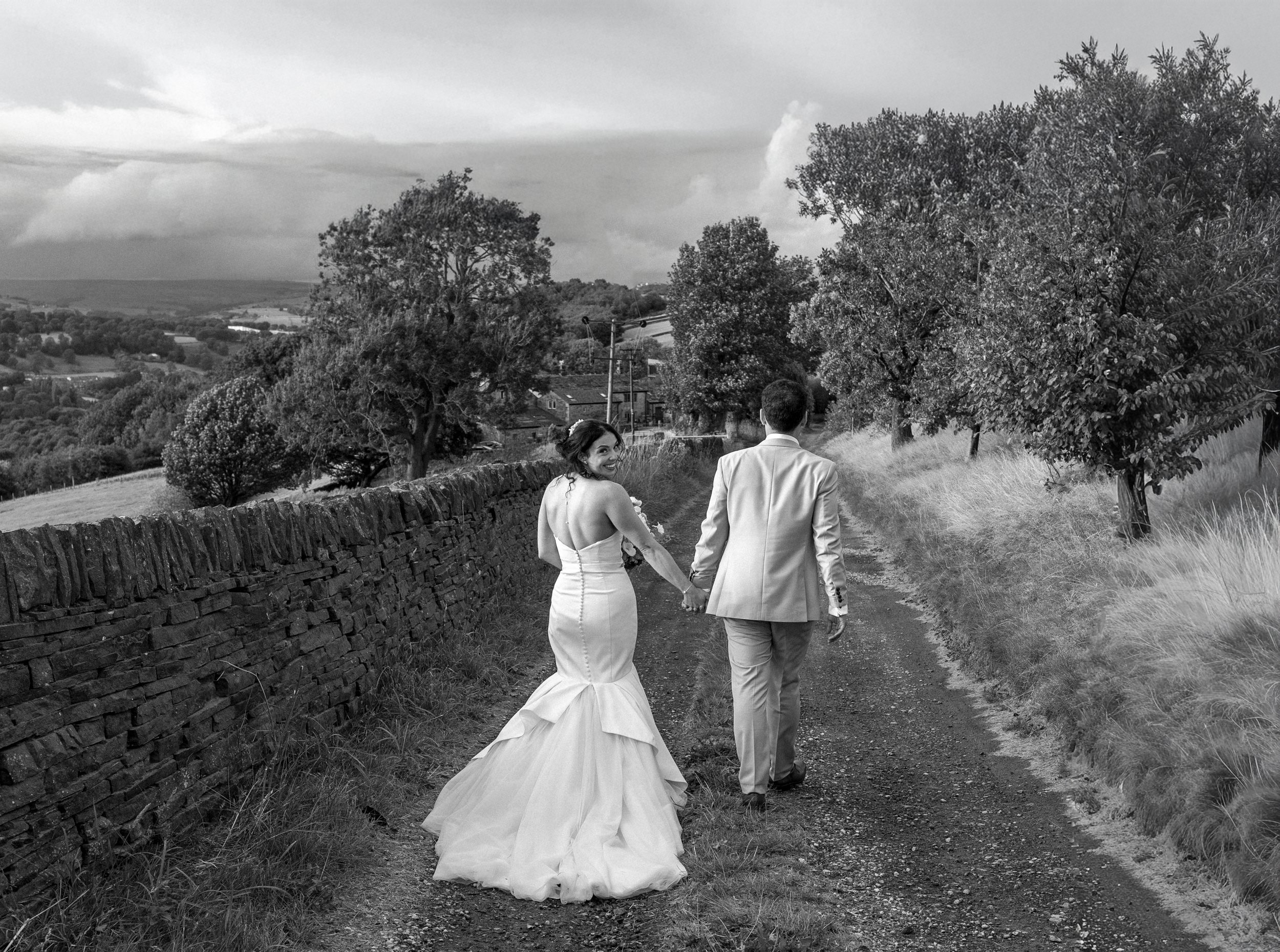 A bride and groom holding hands and walking on a country trail, with trees and landscape in the background, black and white photo.