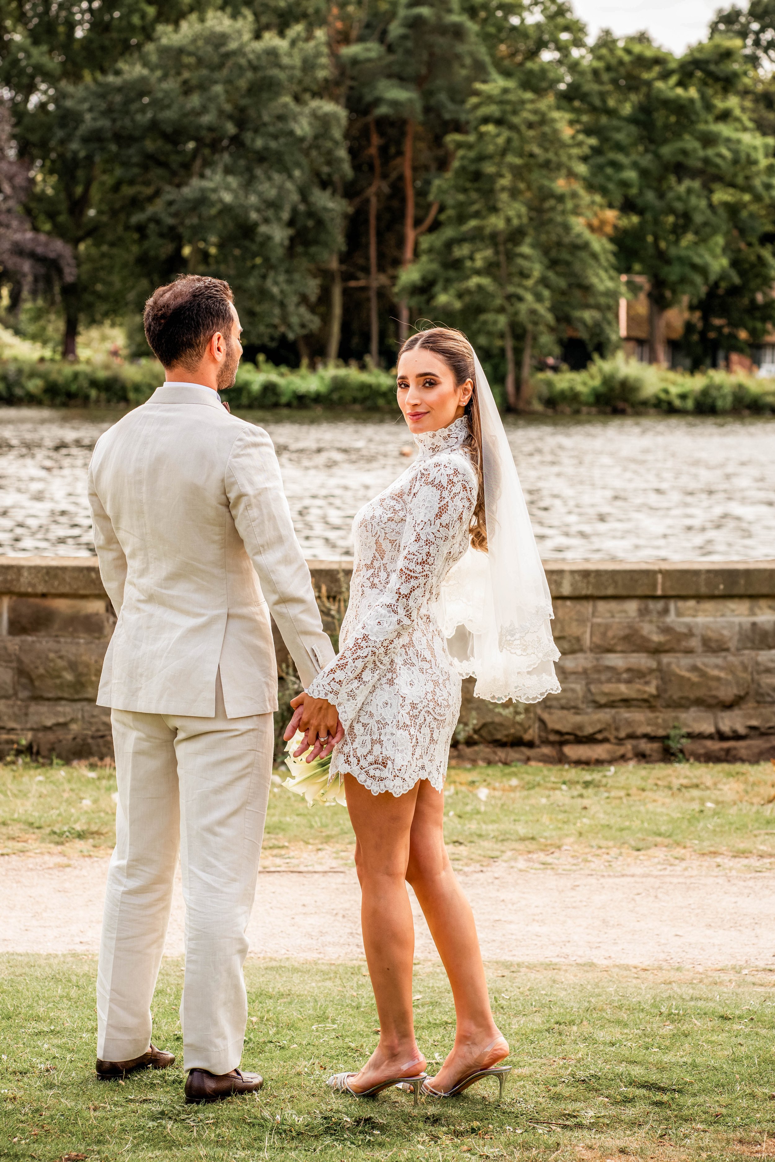 A bride and groom holding hands outdoors beside a river, with trees in the background, during their wedding ceremony.