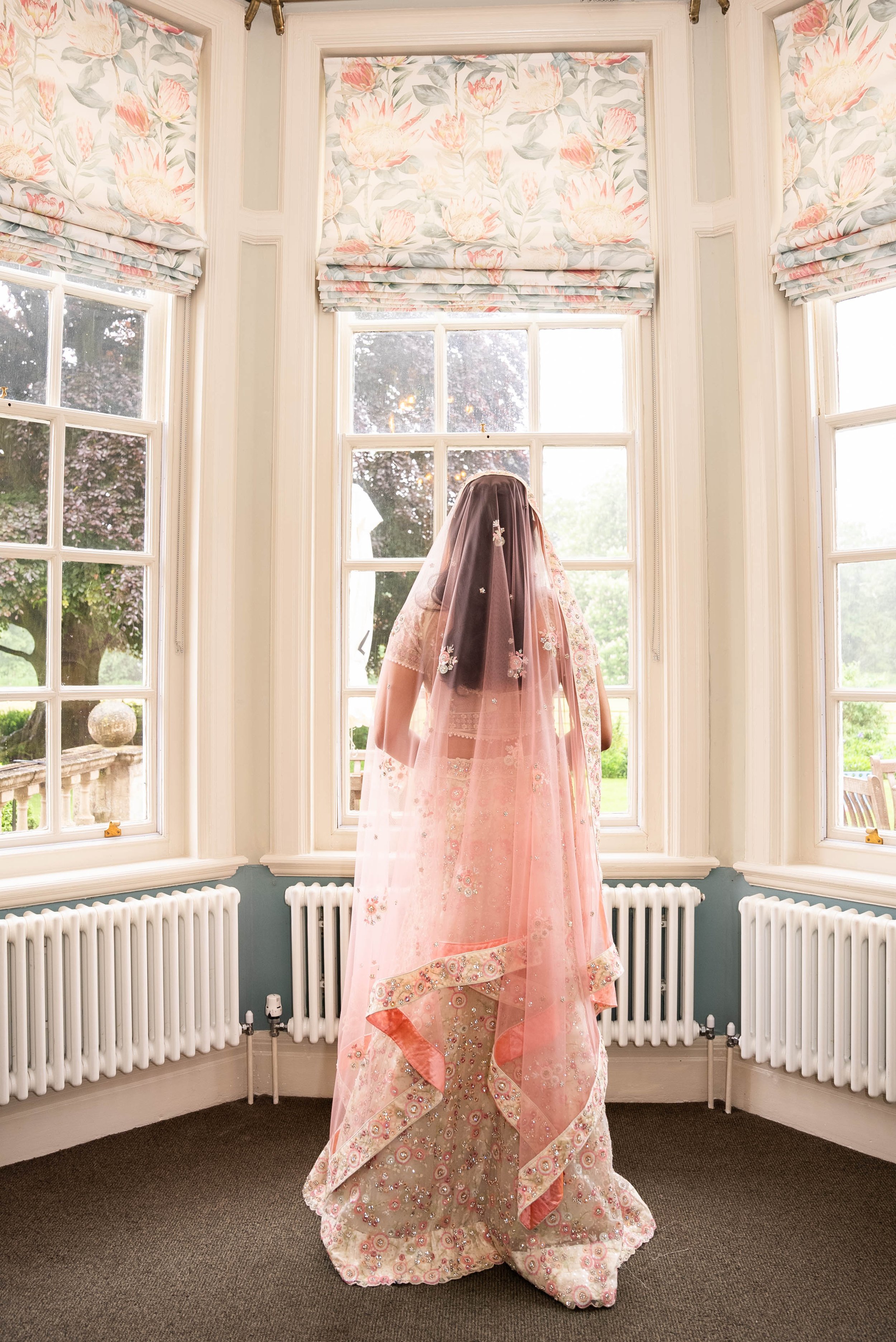 A woman dressed in traditional pink and gold Indian bridal attire standing by a bay window with floral Roman shades, looking outside at a lush garden.