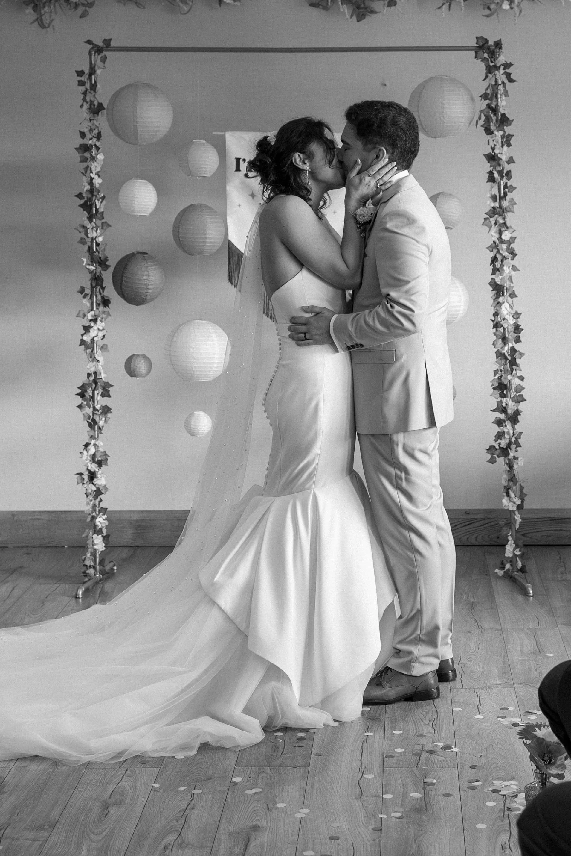A bride and groom sharing a kiss during their wedding ceremony, surrounded by hanging paper lanterns and decorative flowers.