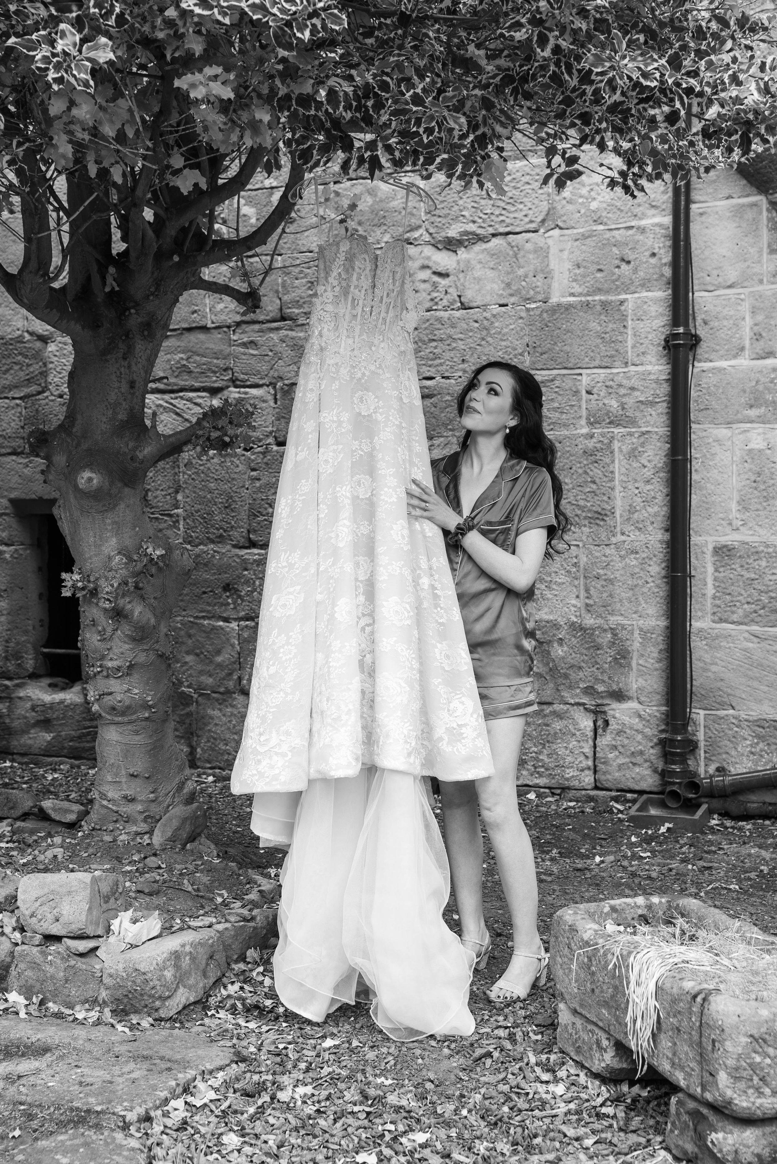 A woman in pajamas looks at a wedding dress hanging from a tree branch in an outdoor setting with a stone wall background.