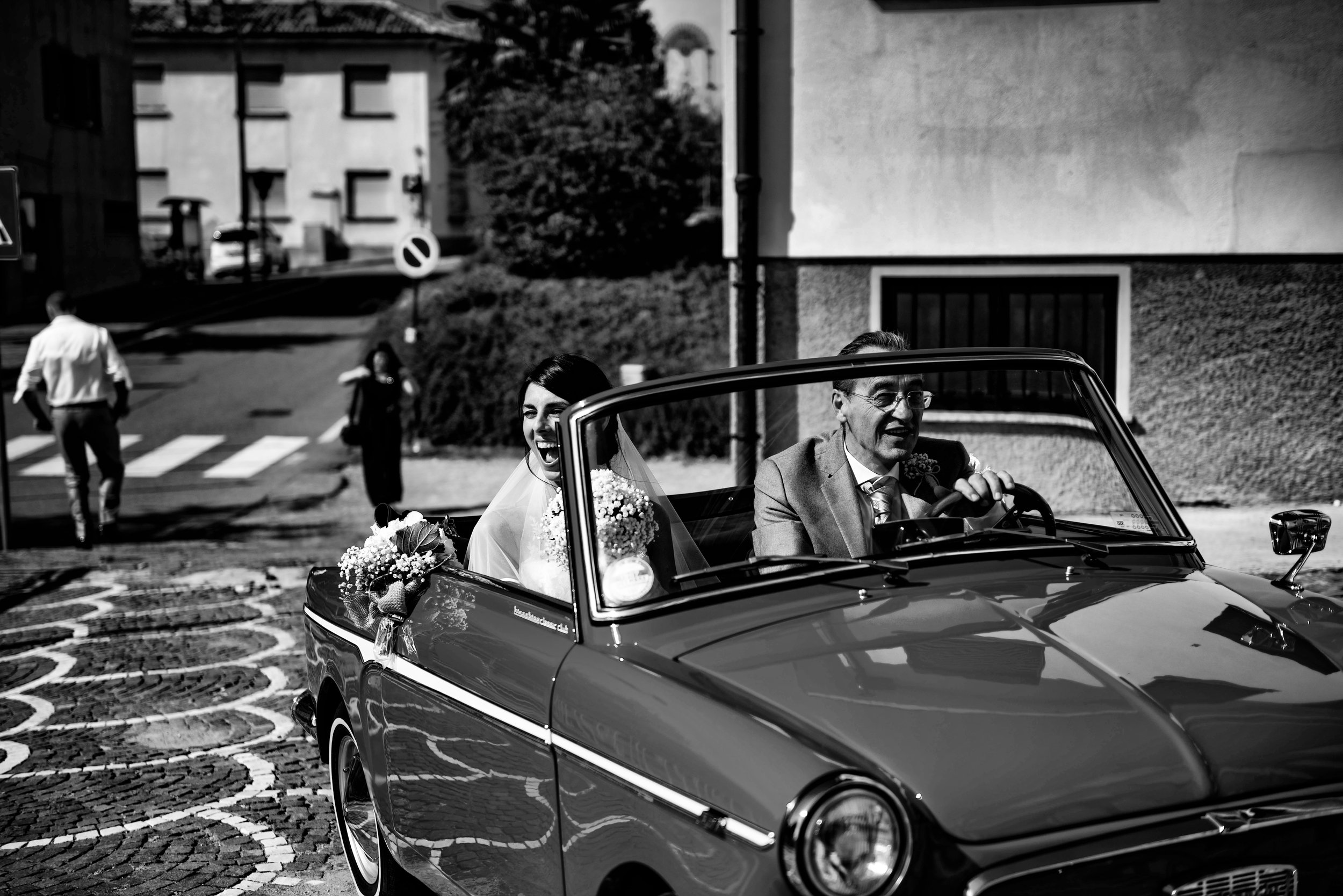 A bride and an older man, possibly her father, riding in a vintage convertible car during a wedding procession on a cobblestone street in a European town. The bride is smiling and holding a bouquet of flowers, with her veil flowing behind her. The ol