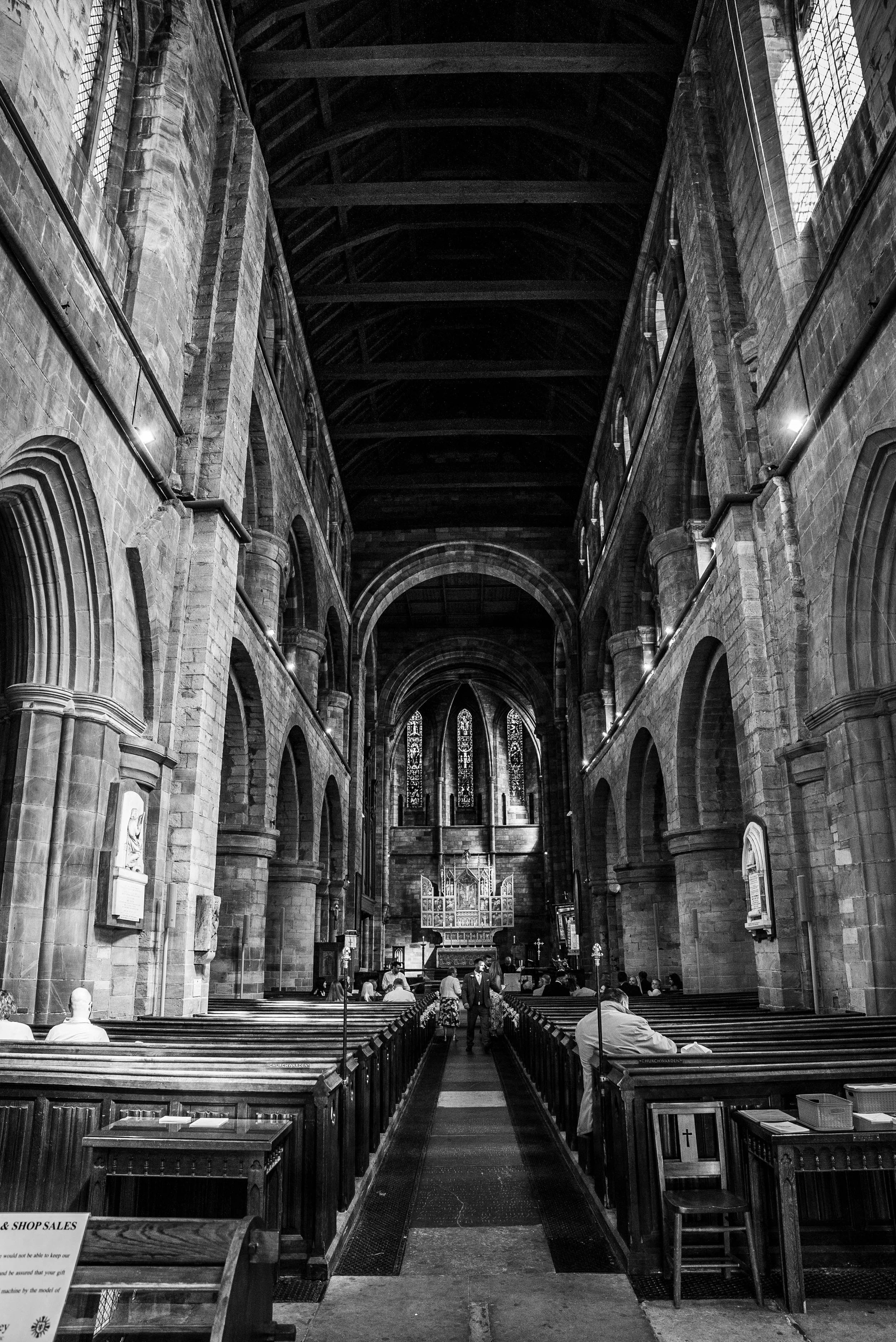 Black and white photo of the interior of a large, historic church or cathedral with high vaulted ceilings, stained glass windows, and wooden pews with some people seated.