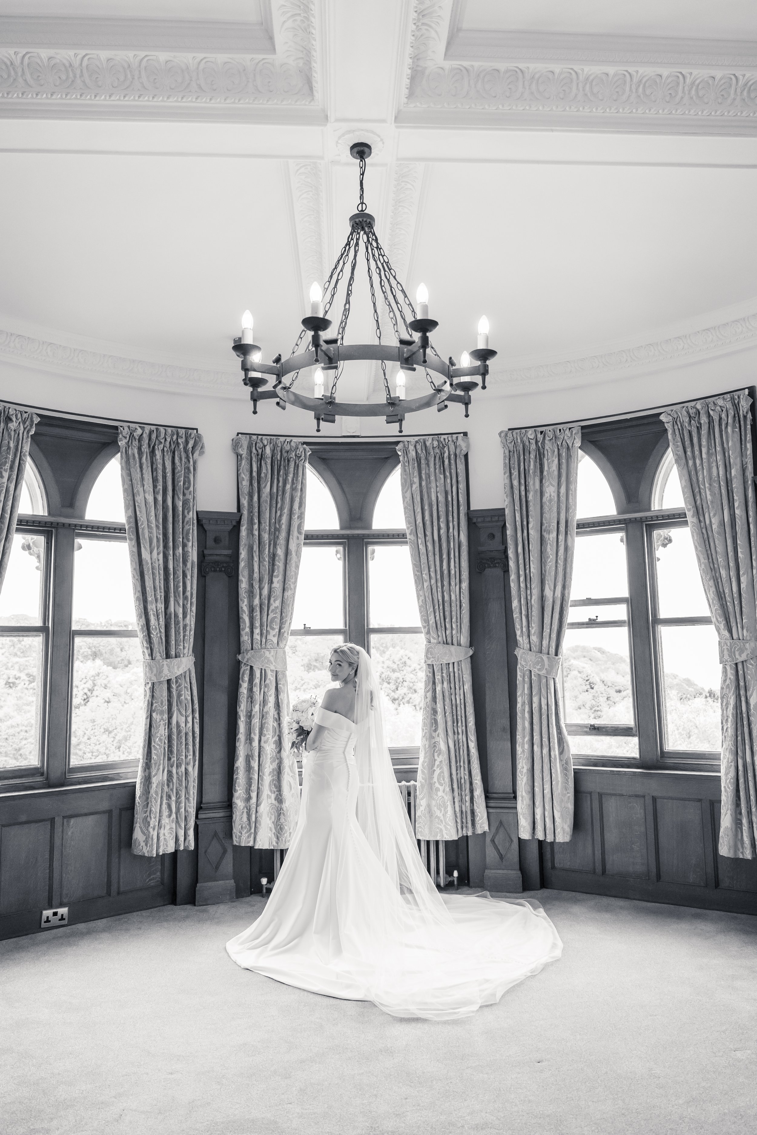 A bride in a white wedding gown standing in a room with large windows and ornate curtains, holding a bouquet, with a large chandelier hanging from a decorative ceiling.