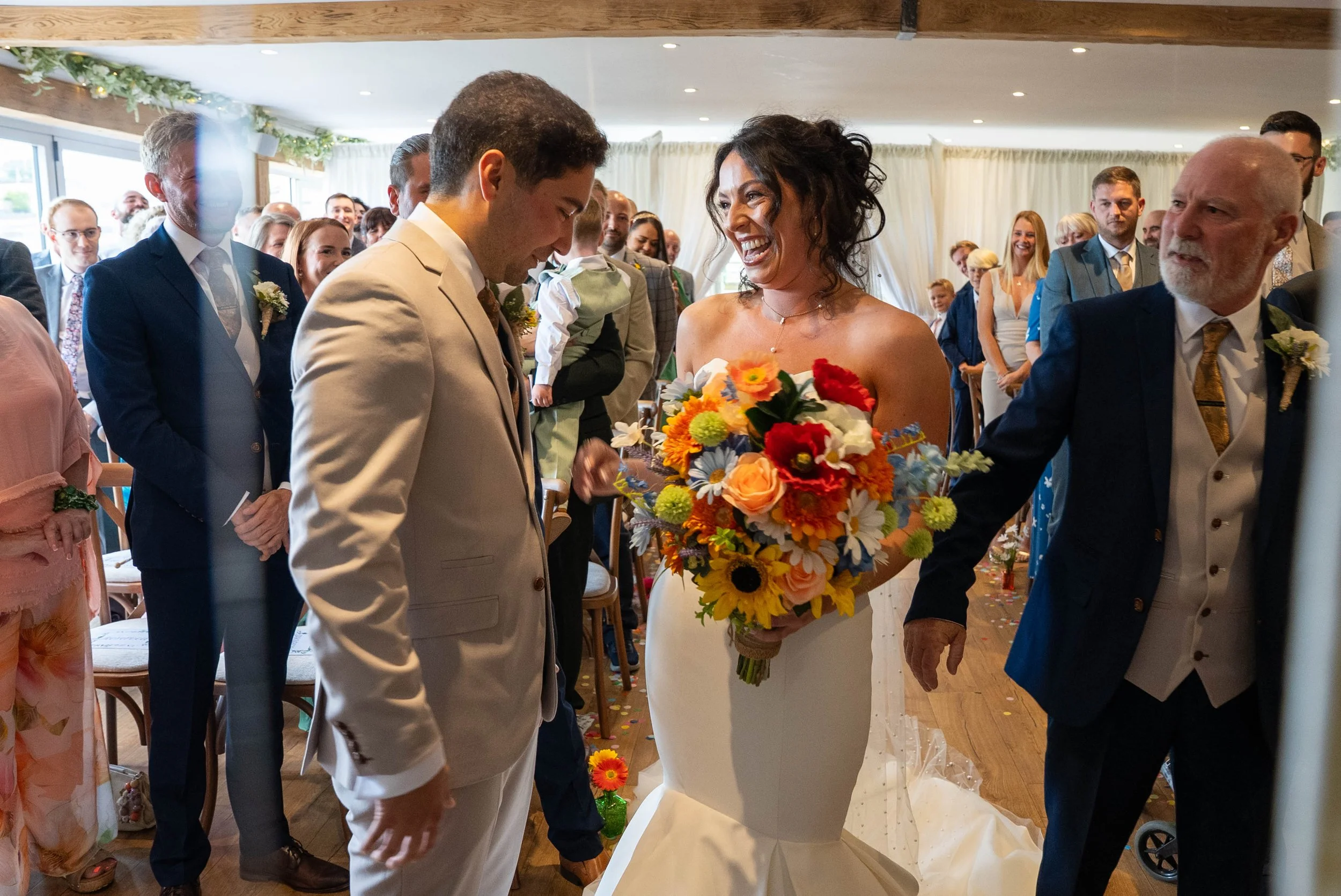Bride holding a colorful bouquet during a wedding ceremony with groom and officiant, surrounded by guests in a decorated indoor venue.